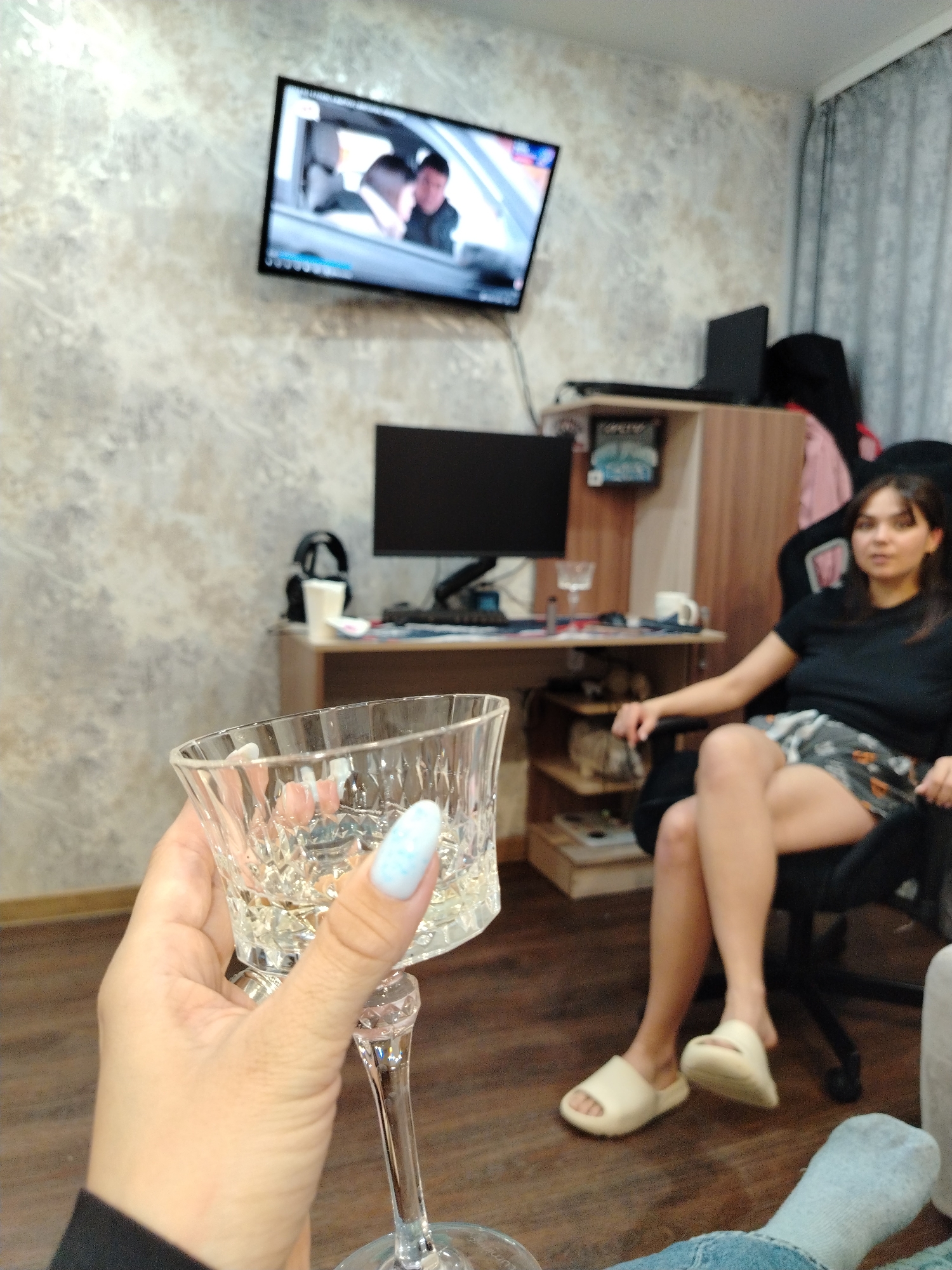 The image captures an indoor scene from a first-person perspective, likely a home in Noviy, Russia. In the foreground, a hand with light blue decorative nails holds a clear, faceted wine glass filled with a light-colored beverage. The person holding the glass is wearing a dark sleeve and part of a leg in a light blue sock is visible at the bottom right.
In the mid-ground, a woman with dark hair is seated in a black gaming or office chair, facing slightly towards the camera. She wears a black t-shirt, patterned shorts, and light-colored slip-on sandals. Her posture appears relaxed, with one leg crossed over the other. Behind her is a cluttered desk setup with a black computer monitor, a keyboard, headphones, and various other items. A black laptop is placed on a shelf above the desk, which also holds some books or boxes.
On the wall above the desk, a television screen is mounted, displaying a scene with two men inside a car, possibly from a video or movie. The wall itself is covered in a textured, light-toned wallpaper. To the right, grey curtains are partially visible, suggesting a window. The floor is made of wood. The overall ambiance is casual and relaxed, typical of an evening at home. No discernible text is clearly visible on any objects or screens.
