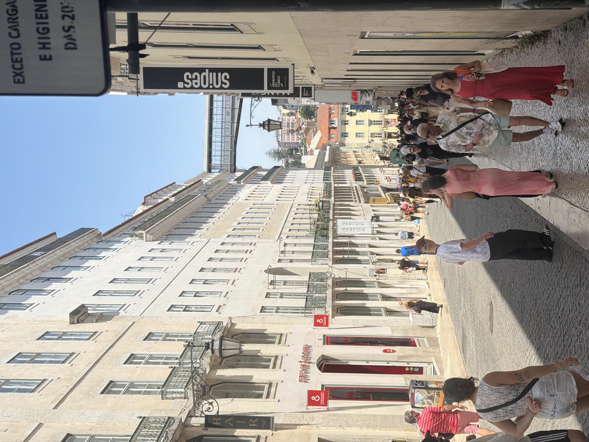 The image depicts a sunny day on Rua Augusta, a pedestrian street in the Baixa district of Lisbon, Portugal. The street is lined with tall, cream-colored buildings with balconies and numerous windows. The cobblestone pavement reflects the sunlight. A diverse crowd of tourists and locals walk down the street, many of them browsing shops.
In the foreground, a woman in a white lace top and white shorts walks away from the camera. To her right, a man in a white patterned short-sleeved shirt and light blue shorts walks towards the camera. To his right, a woman in a long red dress walks towards the camera, holding a water bottle.
Further down the street, a snipes sign is visible. A pedestrian bridge spans the street in the distance. Buildings in the background are a mix of architectural styles, with some featuring red-tiled roofs. The sky is clear and blue. Various shops are visible along the street, including "Perfumes & Companhia" and "H&M". The overall scene is one of a bustling urban environment on a bright, warm day.