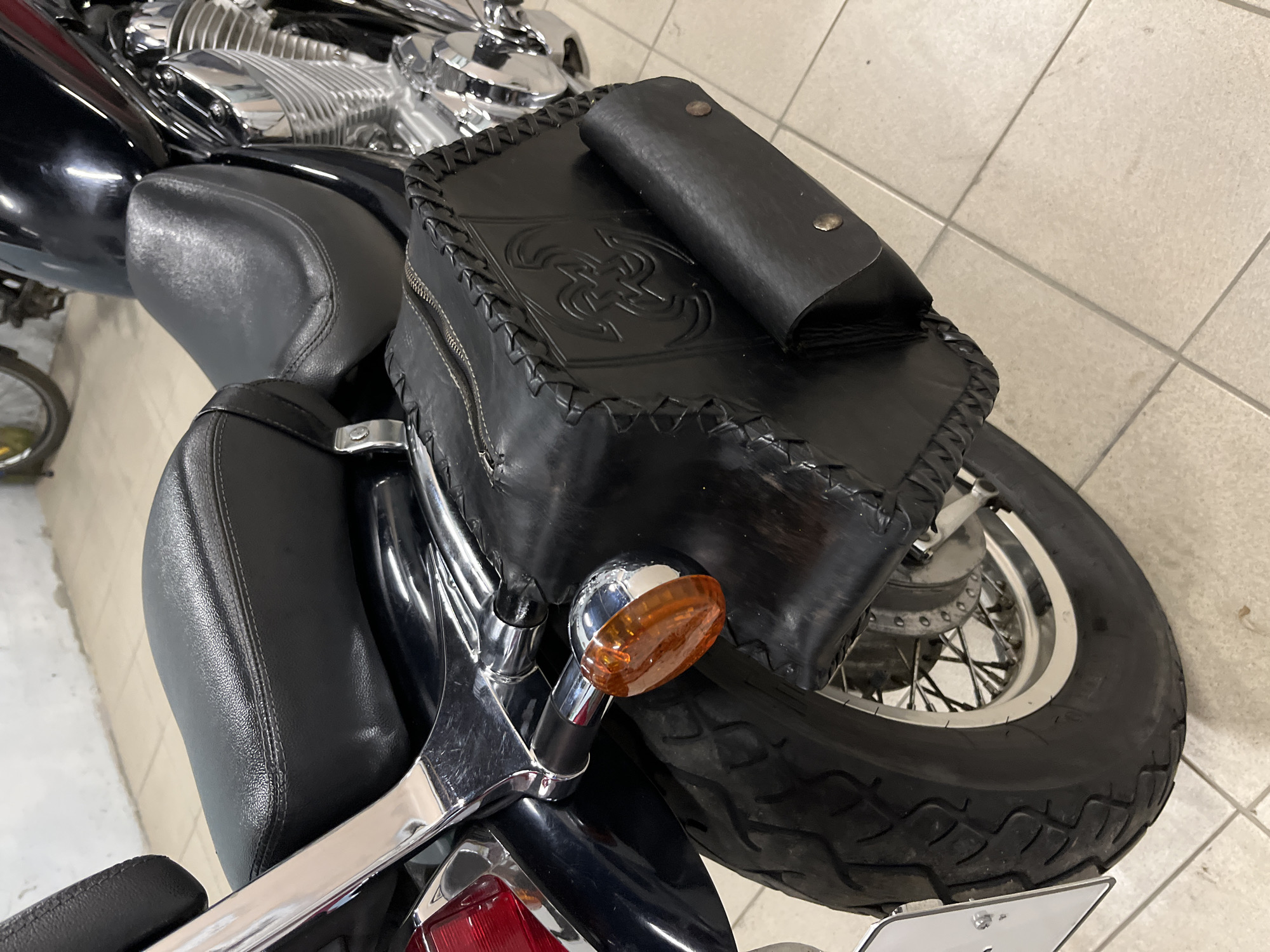 A black leather saddlebag is attached to the rear of a black motorcycle. The saddlebag has decorative braiding around its edges and an embossed design in the center. A smaller flap pocket is also visible on the front of the saddlebag. The motorcycle's rear tire and part of its chrome engine are also visible. The motorcycle is parked on a tiled floor. The background is out of focus but appears to be an indoor space, possibly a garage or workshop. There is no indication of the city of Myrhorod, Ukraine in the image.