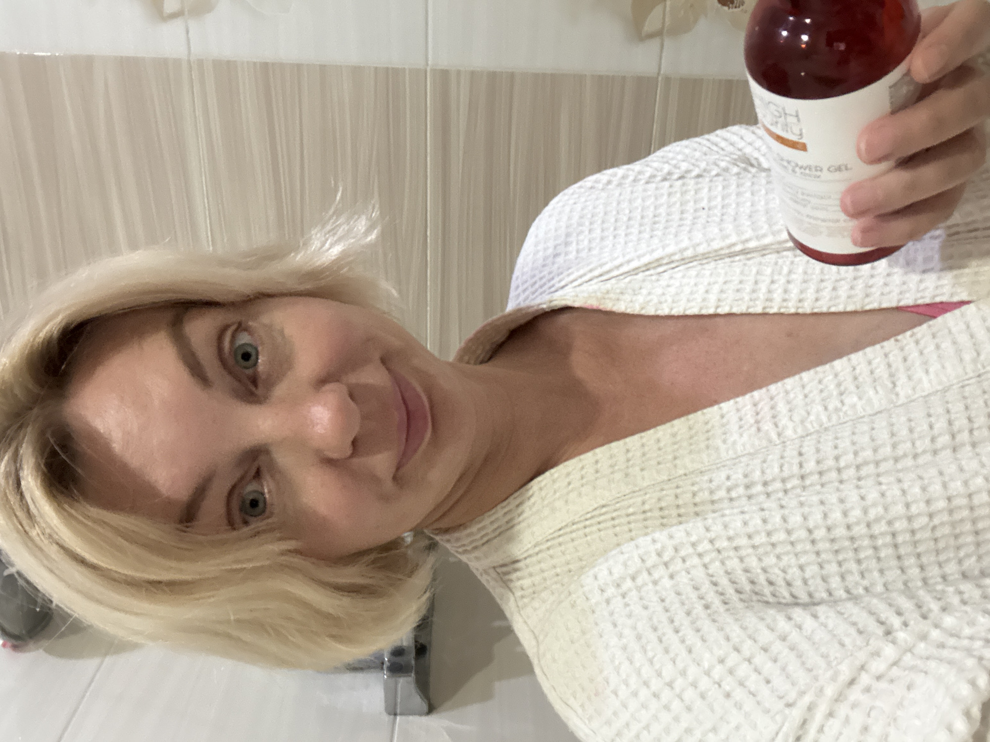A woman with blonde hair, wearing a white waffle-knit bathrobe, holds a bottle of red liquid, possibly shower gel, in her right hand. She is looking at the camera with a slight smile. The background appears to be a bathroom with tiled walls. The image is a selfie, suggesting the woman is taking the photo herself. The location is not specified in the image or context provided.