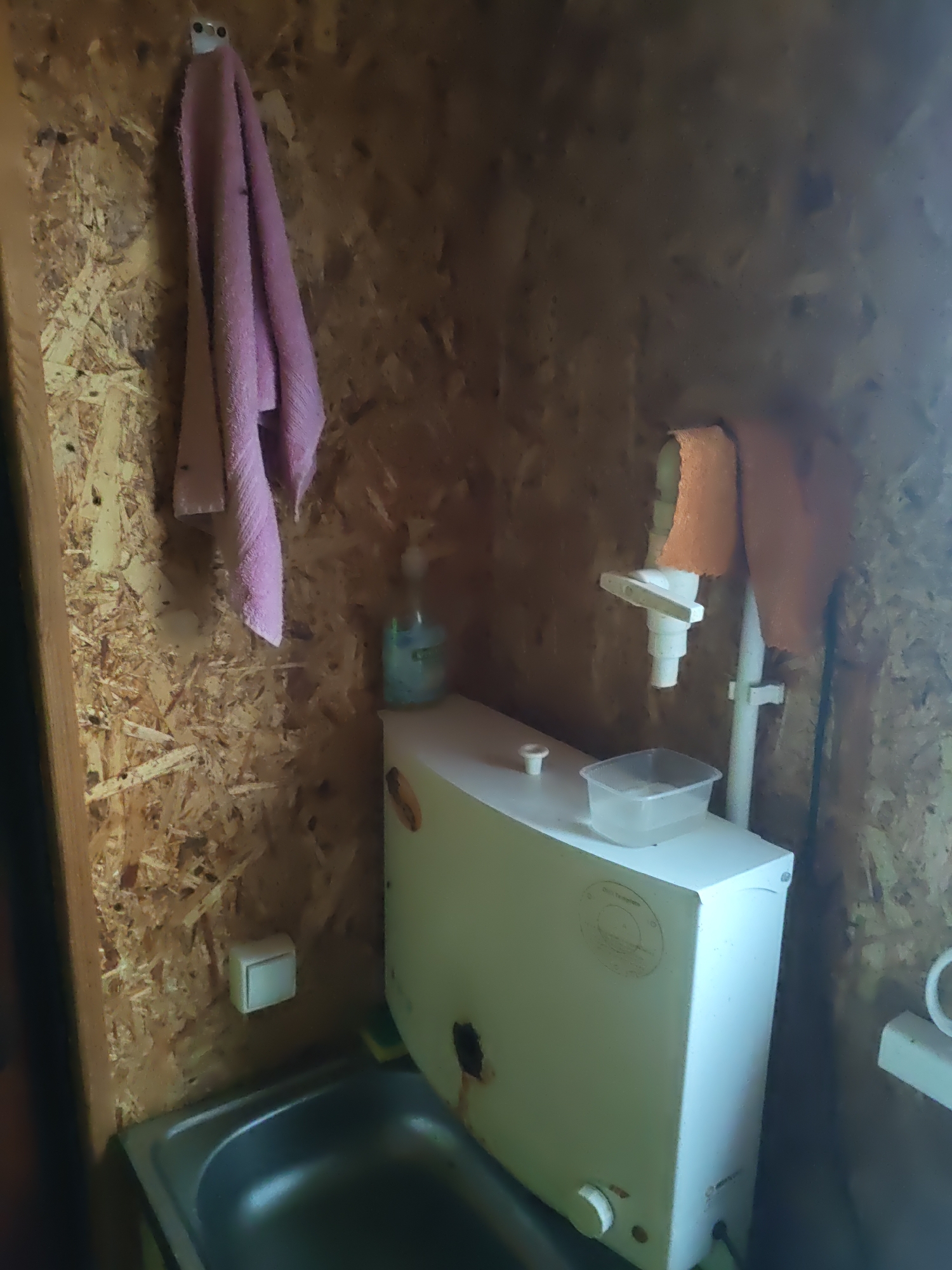 The image shows a small, rustic bathroom. A pink towel hangs on a hook on the left wall. To the right of the towel is a blue plastic bottle, likely containing cleaning fluid. In the center of the image is a white cistern, possibly for a toilet, with a small plastic container on top. A pipe assembly with an orange towel draped over it is visible to the right of the cistern. A stainless steel sink is partially visible in the foreground on the left. The walls appear to be made of particle board or plywood. The overall impression is of a functional but basic facility. The location is identified as Platoshino, Russia, suggesting this is likely a rural or basic dwelling.