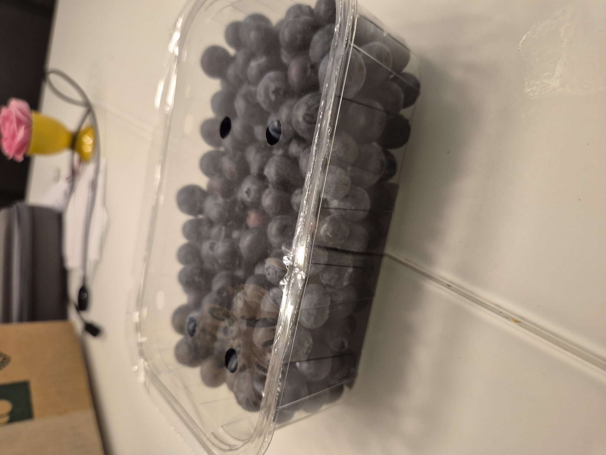 In Ergolding, Germany, an indoor scene depicts a clear rectangular plastic container filled with dark blue to purple blueberries, exhibiting a natural waxy bloom. The container, positioned centrally, features a ridged top edge and at least two visible circular ventilation holes on its translucent lid. It rests on a light-colored, possibly off-white or light grey, tiled surface with discernible grout lines. In the blurred background, to the upper left, a section of a brown cardboard box with a partial dark green marking is visible. Adjacent to it, a dark grey or black electronic device and associated cables are partially present. To the upper right, a yellow goblet-shaped item holds a pink, ruffled object resembling an artificial rose. No individuals or active interactions are visible. The scene is illuminated by artificial light, causing reflections on the plastic container.