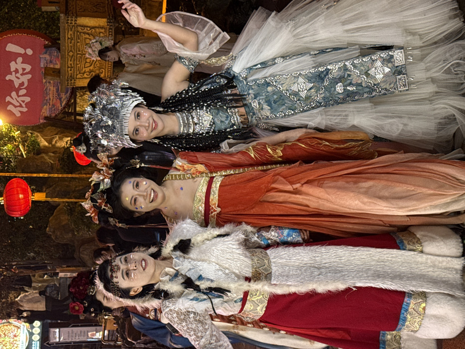 An image taken at night in Yuzhong District, China, shows three adult women, dressed in elaborate traditional-style costumes, posing for a photograph. The women are positioned from left to right, facing the viewer with expressions ranging from a slight smile to a broad grin.
The woman on the far left wears a white, long-sleeved, semi-sheer lace top, covered by a white faux-fur vest. Her lower attire consists of a red skirt with ornate gold and blue trim at the waist and hem. She has decorative white markings on her face.
The central woman is adorned in a flowing, multi-layered gown in shades of orange and red, with gold accents on the bodice and sleeves. Her hair is styled in an intricate updo featuring red and gold floral adornments. Scattered glitter is visible on her face.
The woman on the right wears a prominent silver, metallic headpiece with intricate designs, complemented by long, dark braided hair extensions that fall over her shoulders. She has a blue and silver patterned crop top with sheer white sleeves, exposing her midriff, paired with a matching long sash or apron. Her lower garment is a voluminous, multi-layered white ruffled skirt. Her left arm is raised and bent at the elbow, hand open. Glitter is also visible on her face.
The background is a dimly lit outdoor environment, suggesting a festive street or market. Prominent features include two red lanterns hanging overhead and a large circular sign to the upper right displaying the red Chinese characters "你好!" (Nǐ hǎo!, meaning "Hello!"). Blurred figures of other people and decorative structures, including an ornate golden object, are visible in the distance. The scene is illuminated by artificial warm lighting.
