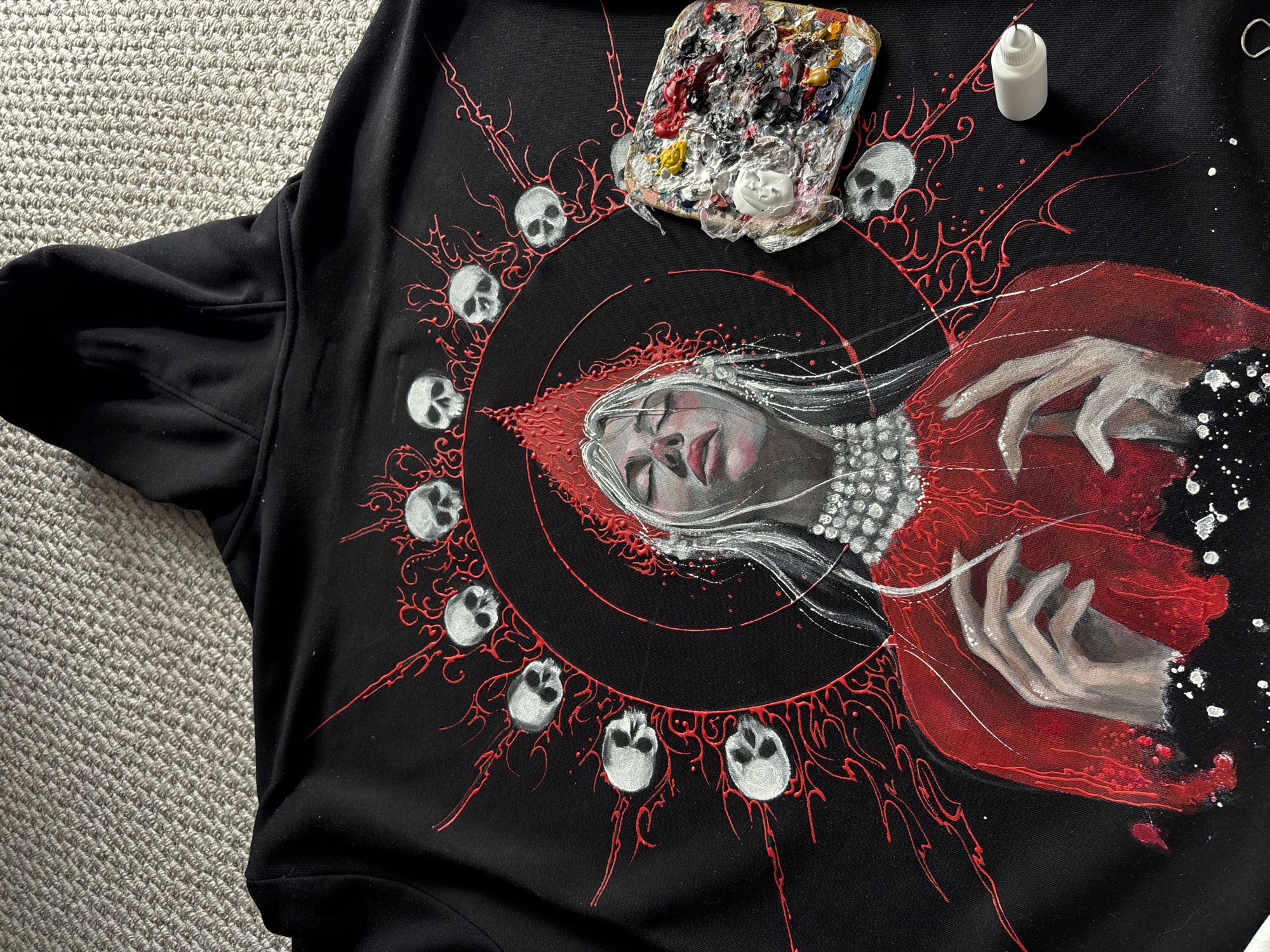 A black fabric item, likely a hoodie or sweatshirt, is laid flat on a light grey, textured carpet. The garment is adorned with an elaborate hand-painted design featuring a central, stylized female figure. Her face is depicted in profile, with eyes closed and white, flowing hair. She wears a rich red garment that envelops her and extends to her visible hands, which are gracefully posed. The figure's skin tone is rendered in shades of grey and white.
Surrounding the figure is a circular design in black, from which intricate, raised red patterns resembling organic tendrils or veins radiate outwards. These red elements connect to and encircle numerous small, stylized white skulls that are arranged in a circular formation around the central figure. White paint splatters are visible on the right side of the artwork, adding a dynamic, textured effect.
In the upper right corner of the fabric, a painter's palette, heavily laden with various colors of dried and fresh paint (including red, white, yellow, black, and blue), is placed. Beside the palette, a small white plastic bottle with a dropper tip, possibly containing paint or a medium, stands upright. The detailed artwork, along with the presence of painting tools, suggests that the piece is either a work in progress or has been recently completed. The overall aesthetic is gothic, mystical, and evokes themes of life, death, and perhaps a fantastical narrative. The location context is Krasnoyarsk, Russia.