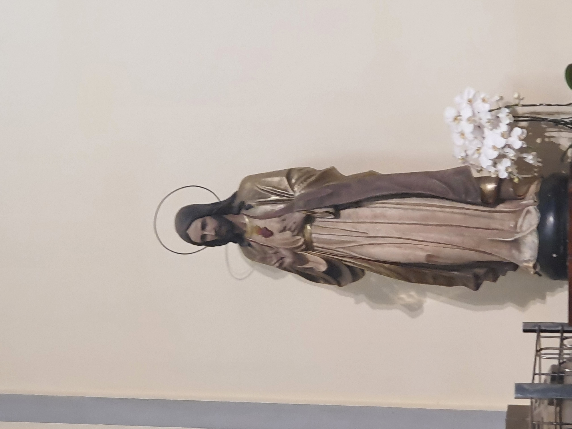 A full-body statue of Jesus Christ stands against a plain, light-colored wall. The statue is depicted with a halo, a beard, and robes, and is holding his right hand up in a blessing gesture and his left hand on his chest, where a red heart is visible. A bouquet of white orchids is placed to the right of the statue. The statue appears to be located indoors, possibly in a church or religious building, with a metal railing visible in the foreground. The image does not provide any information about its specific location in Central Jakarta, Indonesia.