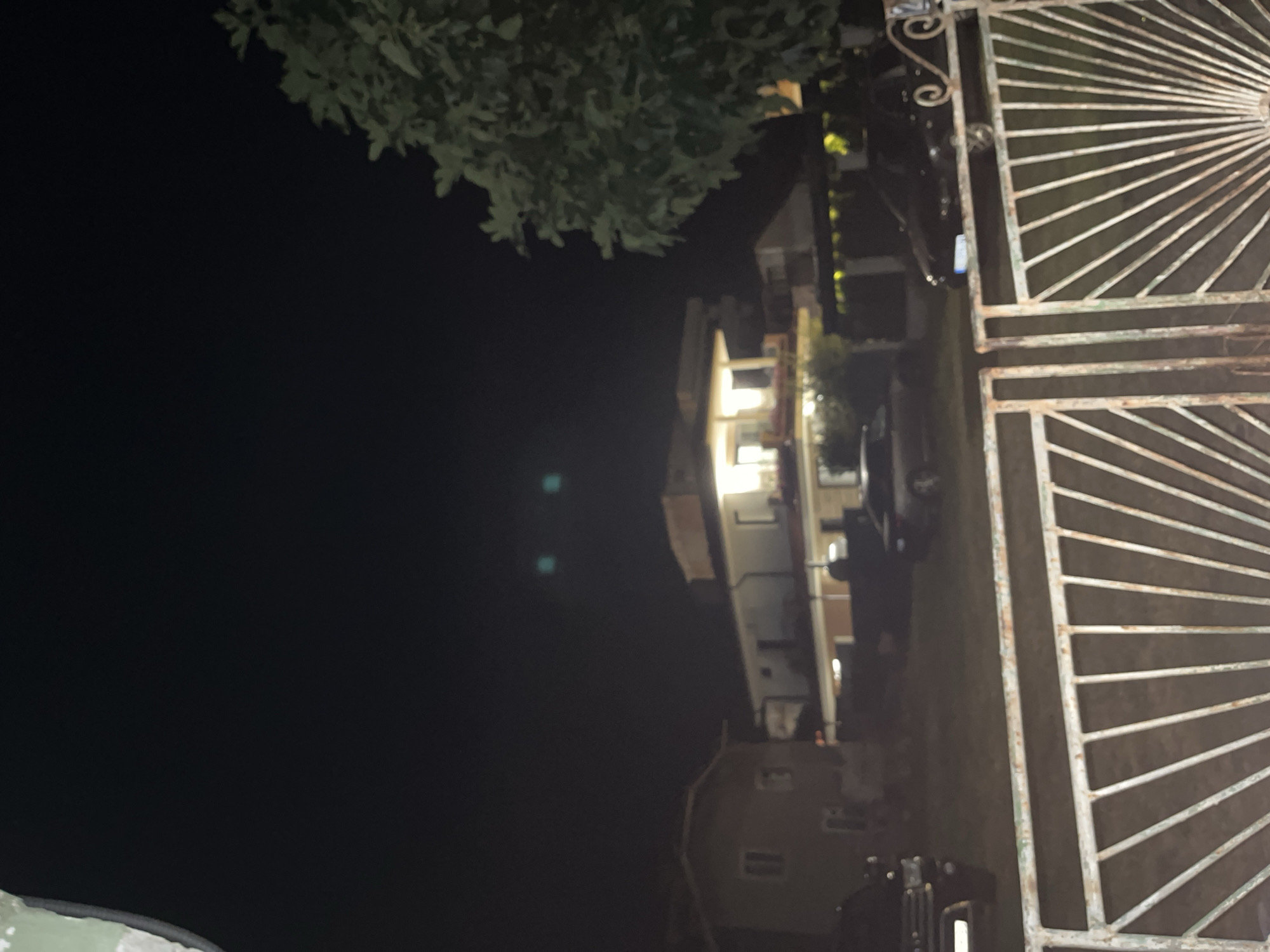 The image depicts a nighttime scene in Obzor, Bulgaria, featuring a residential area. In the foreground, a rusted white metal gate with a sunburst design is prominent. Beyond the gate, a grassy yard is visible, with several vehicles parked. To the left, a dark-colored SUV is partially visible, and to the right, a black sedan is parked. In the background, two houses are illuminated. The house on the left appears to be a lighter color with visible windows. The house on the right is a multi-story structure with bright lights illuminating its windows and facade. A large tree with dense foliage is situated to the right side of the frame, extending from the top edge. The sky is dark, with two indistinct light sources, possibly streetlights or reflections, visible in the upper middle portion of the image. The overall impression is of a quiet residential street at night.