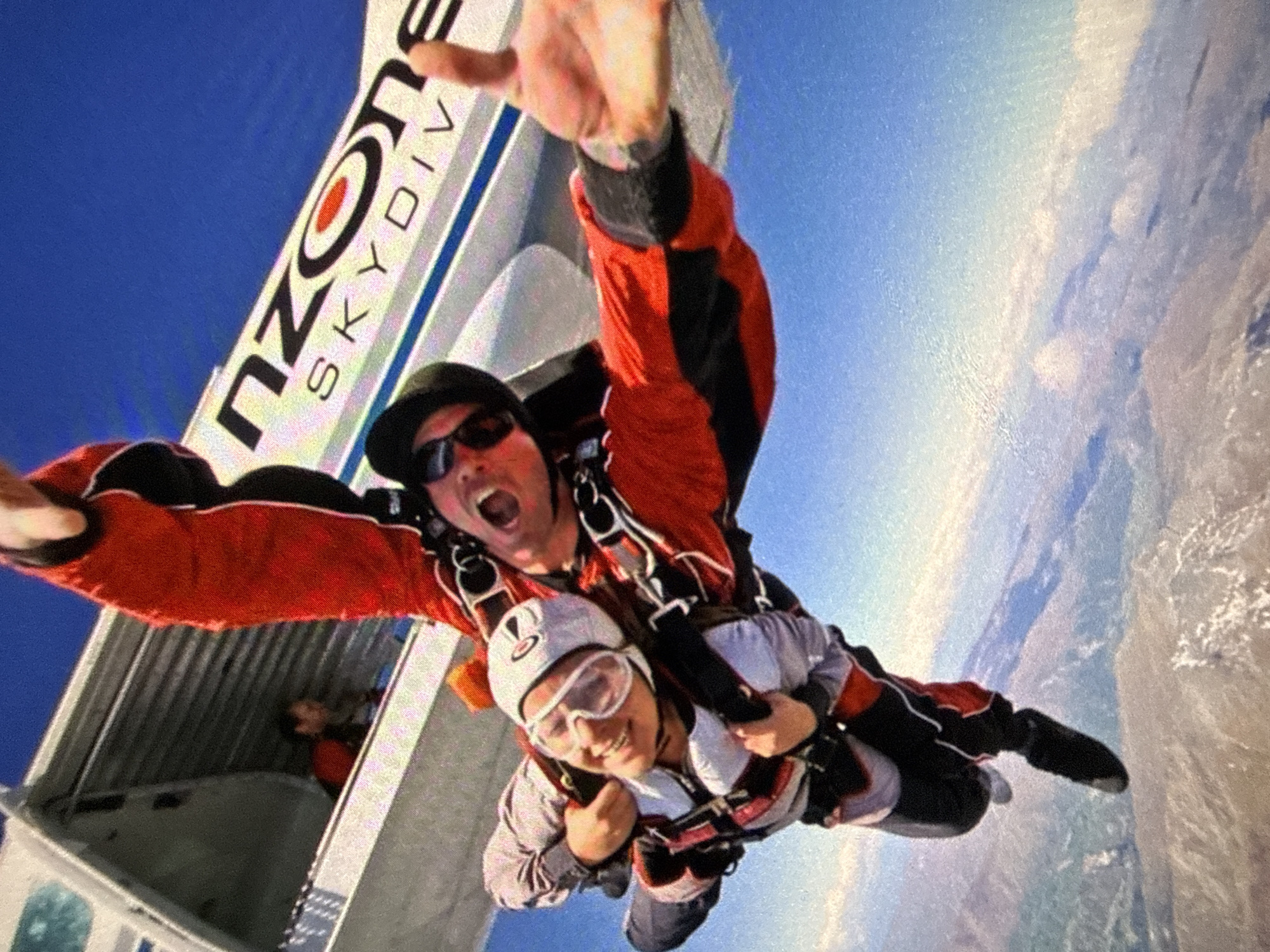 Two individuals are skydiving, tandem, from an airplane. The person in the front wears a helmet and goggles and is smiling. The person in the back wears a hat and sunglasses and has their mouth open, yelling. The plane reads "nZONE SKYDIV." The background shows the sky and land. This image was taken in Queenstown, New Zealand.