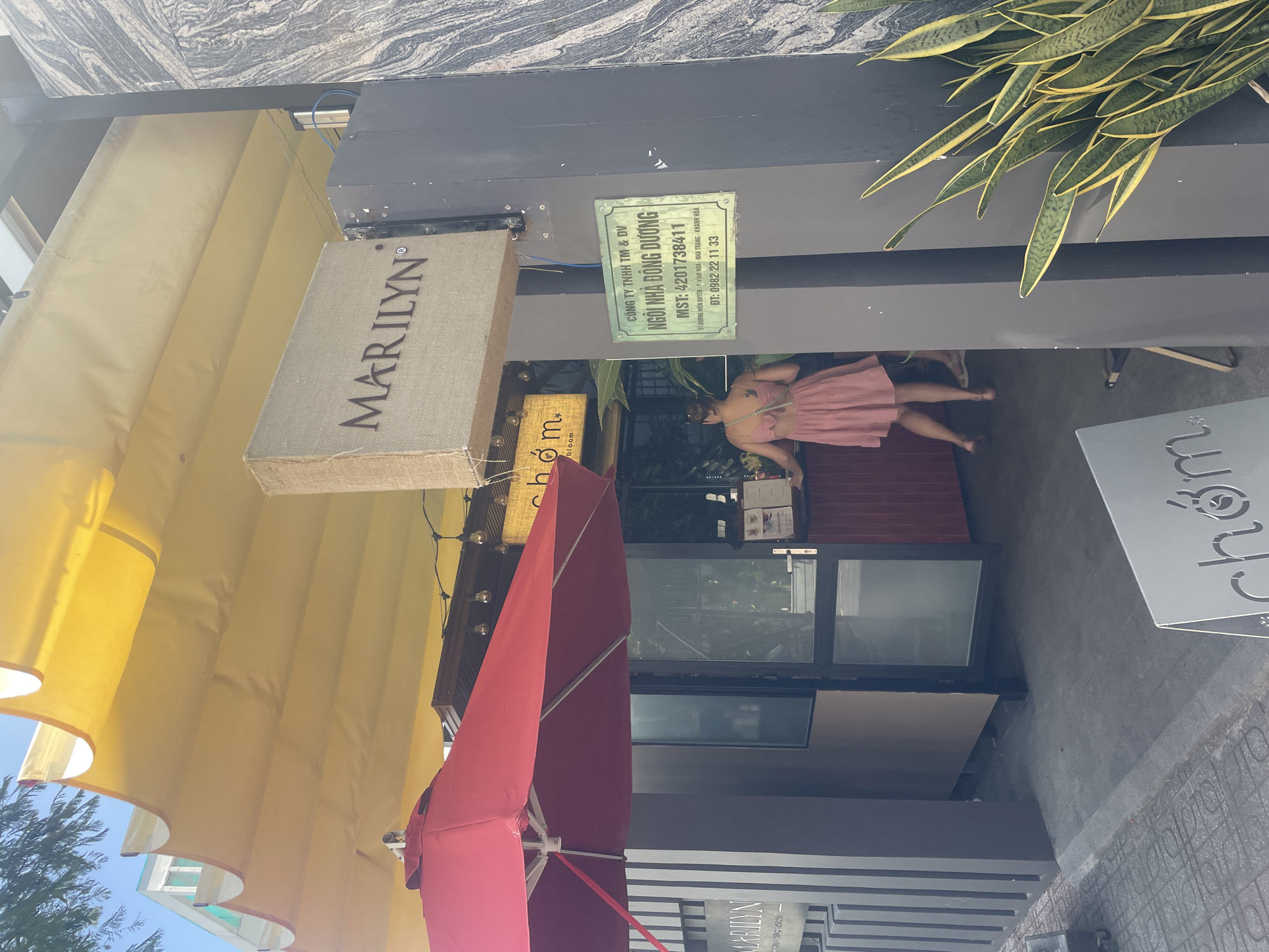 This image depicts the exterior of a commercial establishment located at 51 Hien Quyen Street, Vinh Hai Ward, Nha Trang, Khanh Hoa, Vietnam, as indicated by a prominent green sign. The establishment operates under the names "NGÔI NHÀ ĐÔNG DƯƠNG" (Indochina House) and also features signage for "MARILYN®" and "chóm bloom."
The scene is set on a bright, sunny day, with clear blue sky visible in the upper left corner through tree foliage. A large, yellow, accordion-style retractable awning extends over the entrance and an outdoor area, providing shade. Below the awning, a large red outdoor umbrella is also open. The building's facade consists of dark grey or black vertical panels and pillars.
One person is visible inside the entrance area, a woman with her hair pulled back, wearing a light pink, backless dress with a bow detail. A small tattoo is discernible on her upper back. She is positioned facing away from the camera, near a dark red counter inside the establishment. The lower legs and feet of a second individual in light-colored footwear are partially visible further inside, behind the first woman.
The entrance features an open glass door. A string of decorative Edison-style light bulbs hangs under the yellow awning. On the right side of the entrance, a large potted Sansevieria plant with green and yellow variegated leaves stands against a dark wall with a grey and white marble-like pattern. An A-frame sidewalk sign, partially visible in the bottom right, displays the "chóm" logo. The ground outside is a paved sidewalk, transitioning to a smooth darker surface at the entrance.