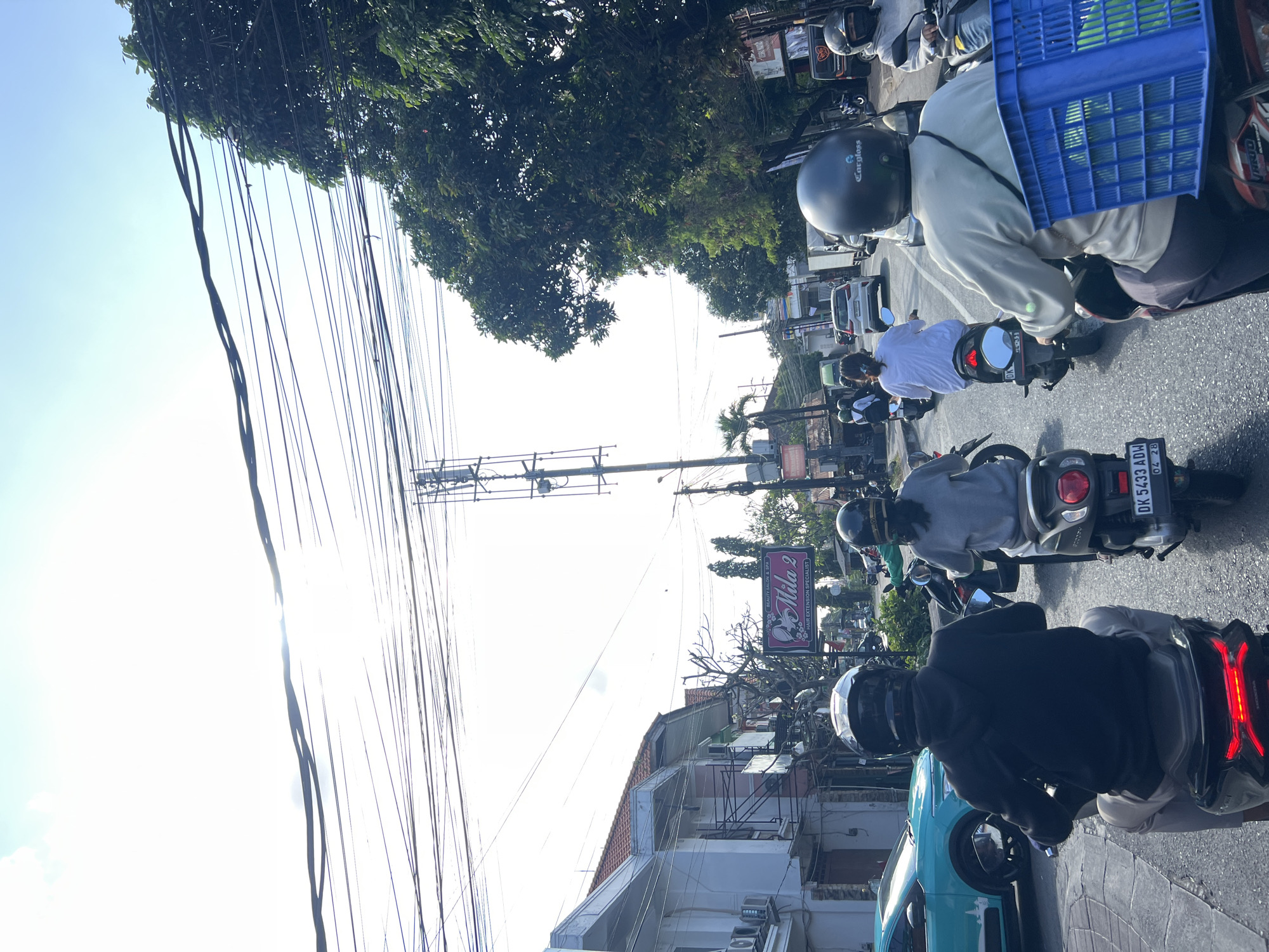 The image depicts a street scene in Denpasar, Indonesia, on a sunny day with a bright sky. The foreground is dominated by several motorcycles and their riders, viewed from behind. The closest rider wears a black hoodie and light-colored pants, with a black helmet. To their right, another rider wears a light grey long-sleeved shirt and dark pants, also with a black helmet. A blue crate filled with green items is strapped to the back of this second rider's motorcycle. Further ahead, other motorcyclists in helmets are visible, some carrying passengers. On the left side of the road, buildings with white and pink facades, air conditioning units, and a sign for "Mila 2 Beauty Salon & Spa Hair Extension Specialist" are present. A turquoise car is parked on the left. The road is lined with trees and utility poles with numerous overhead wires crisscrossing the sky. In the distance, more vehicles and buildings can be seen. The overall impression is that of a bustling urban environment during daylight hours.