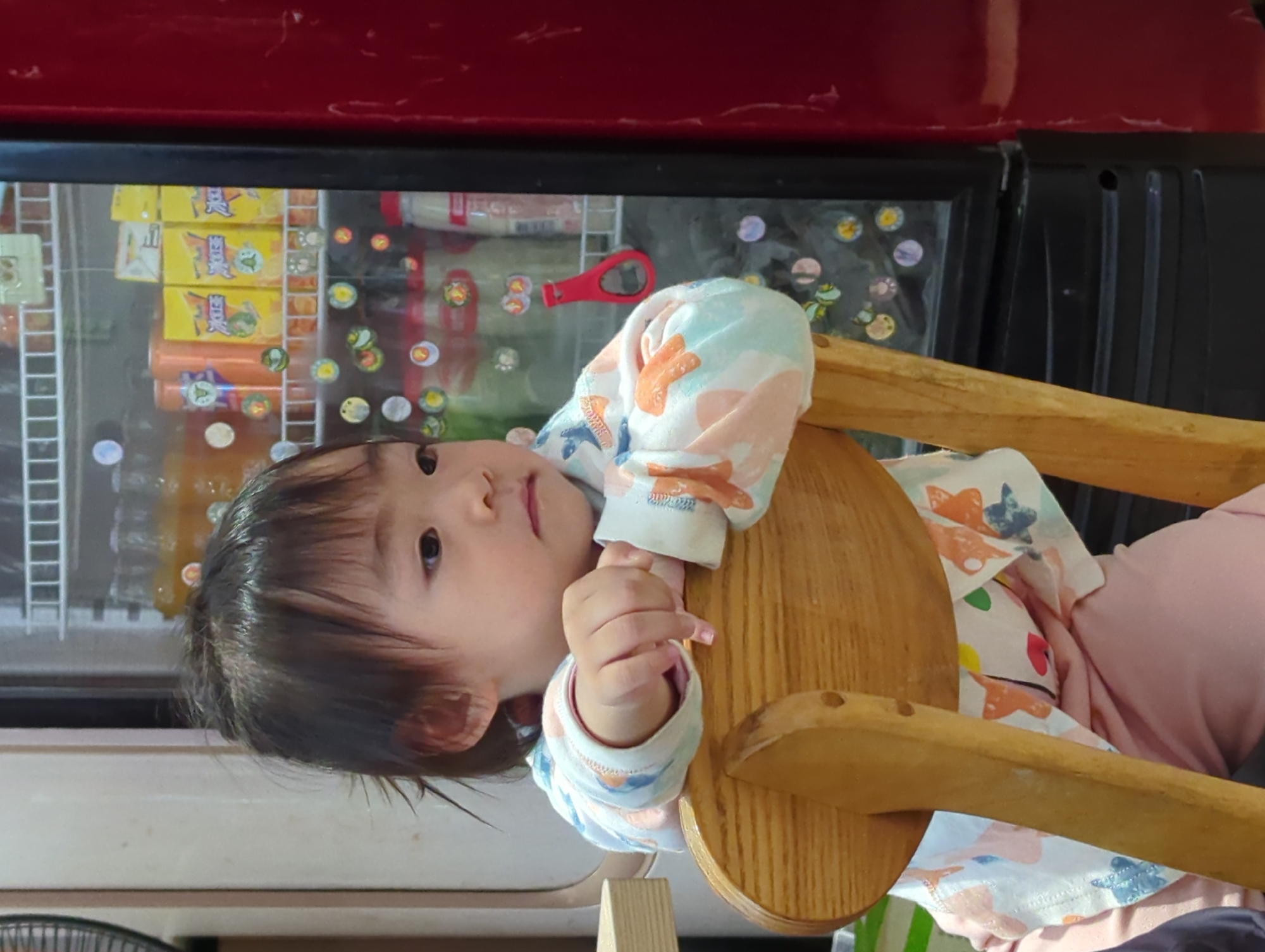 A toddler with dark hair is sitting in a wooden chair with a rounded back. The child is wearing a long-sleeved shirt with light-colored patterns, and light pink pants. The toddler is facing left with a slightly downward gaze. The background includes a refrigerated display case with various items inside, some covered with stickers. The top of the case is red. The location appears to be a store in the city of Shunde District, China.