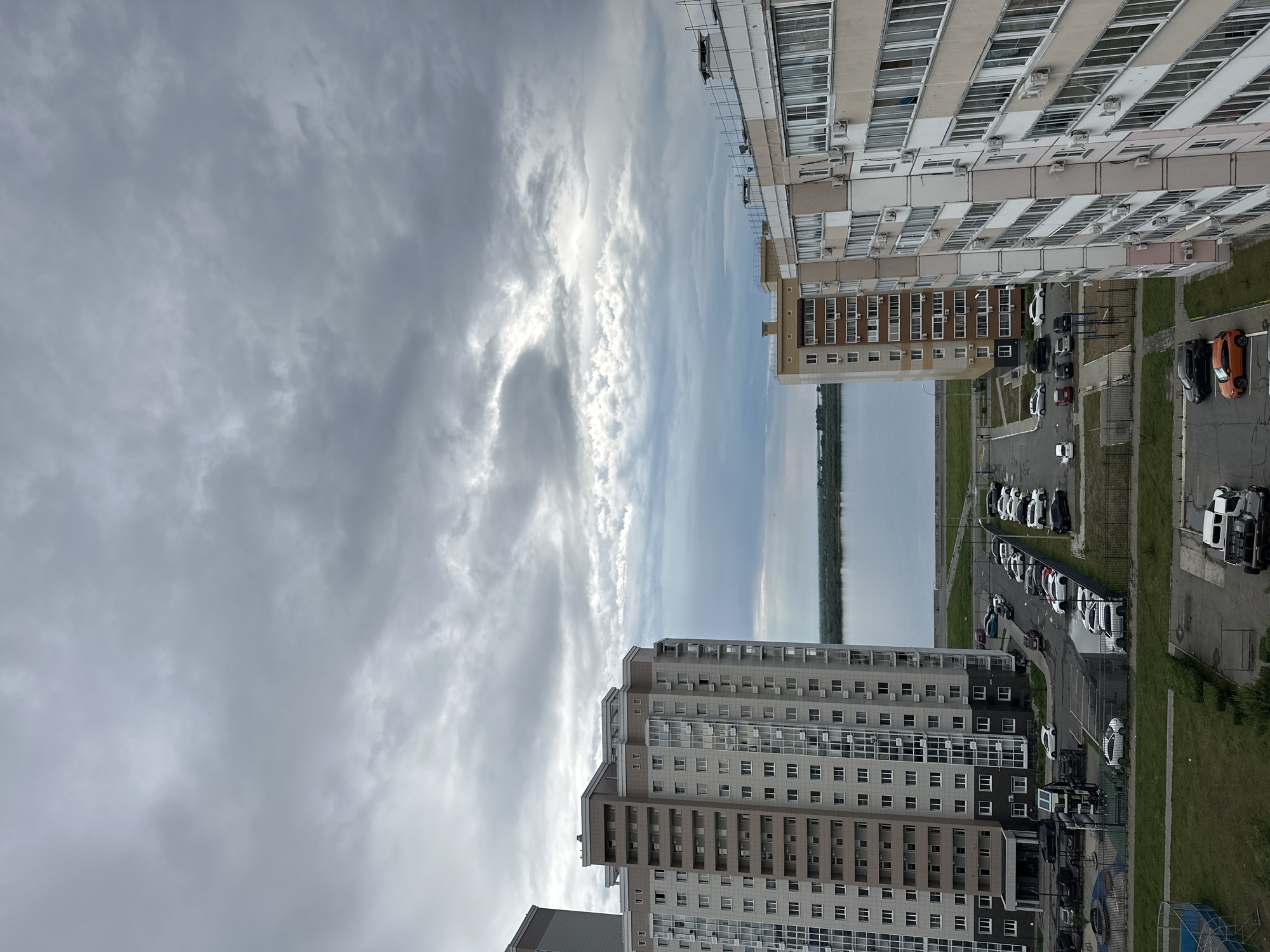 The image depicts a cloudy day in Khabarovsk, Russia, overlooking a wide body of water, likely the Amur River, which forms the border with China. In the foreground, several modern apartment buildings are visible. The building on the left is tall with a light brown and cream facade, featuring numerous windows and balconies. The building on the right is also a multi-story apartment complex, with a lighter cream and beige exterior, also heavily windowed. Between the buildings, a parking lot is visible with several cars parked, including white, black, and orange vehicles. A playground with blue and yellow structures is located in the lower left portion of the image. The sky is overcast with dramatic, layered gray clouds. The water body stretches across the horizon, with a distant treeline on the opposite bank. The overall scene conveys a sense of urban residential life under a dramatic sky.