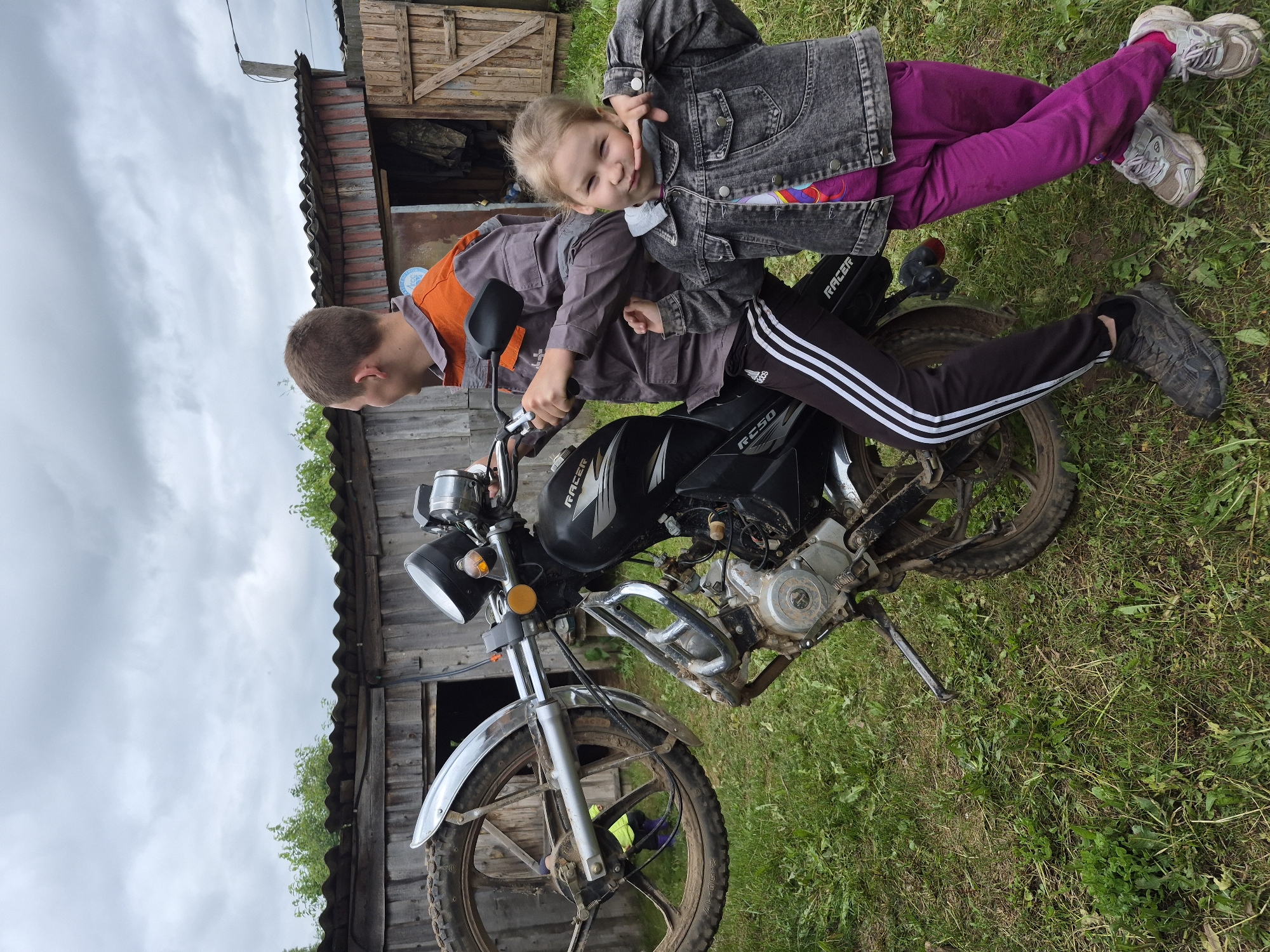 In the city of Kharnavy, Russia, an outdoor scene on an overcast day features a young adult male and a young girl with a black Racer RC50 motorcycle. The male, wearing a grey and orange work jacket, black Adidas track pants with white stripes, and muddy dark sneakers, is seated on the motorcycle, facing left and away from the camera. The motorcycle's front wheel assembly, detached from the forks, is held vertically to the left, displaying a muddy tire and chrome fender. A young girl, approximately 5-8 years old, with light hair, dressed in a dark denim jacket, a colorful t-shirt, bright pink pants, and dirty light-colored sneakers, stands to the right of the male, facing the camera with a playful expression, holding her finger to her chin. The immediate ground is covered with overgrown grass and weeds. In the background, a weathered wooden shed with a dark corrugated roof features a blue circular decal on its wall. One shed door is open, revealing a partially visible individual in a yellow-green top bending inside. Green foliage is visible above the shed, and the sky is uniformly overcast grey.