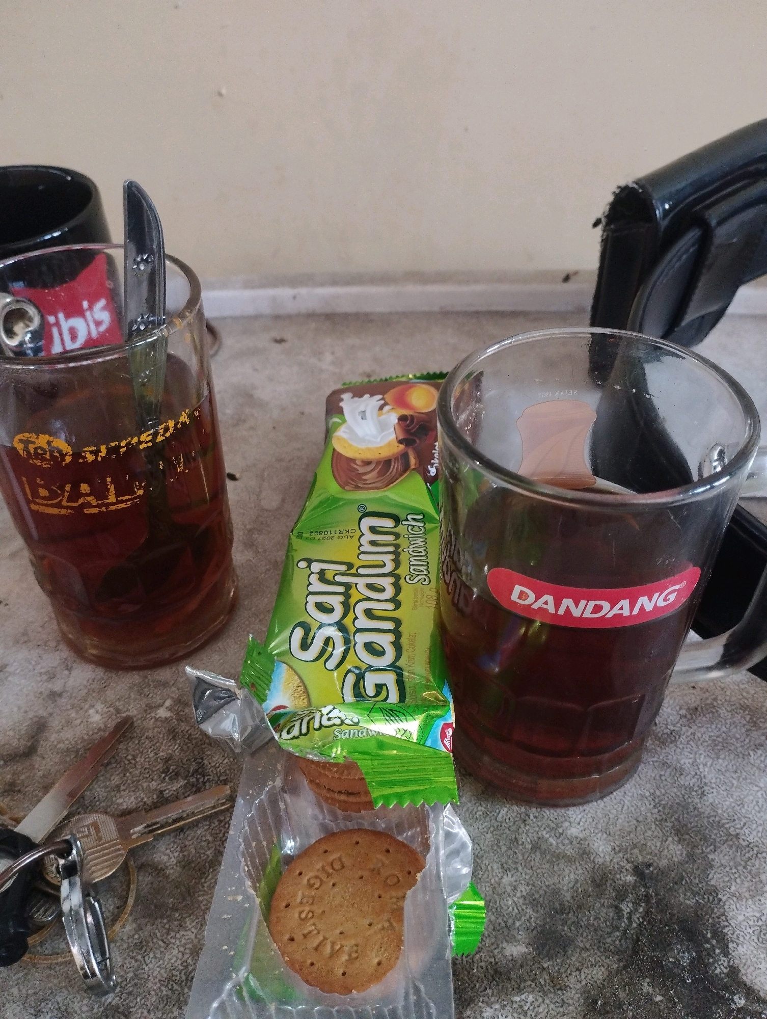 This is a close-up, eye-level, still shot of two glasses of iced tea, a package of biscuits, and a set of keys on a grey textured surface. The lighting is dim, and the background is a plain, light-colored wall. In the foreground, to the left, is a glass of iced tea with a red and white label that reads "ibis". A silver spoon is resting in the glass. To the right, another glass of iced tea has a red and white oval sticker that reads "DANDANG". Between the glasses is a green package of "Sari Gandum Sandwich" biscuits. To the left of the glasses, a set of keys and a small knife are lying on the surface. A black mug is partially visible behind the left glass of iced tea. The overall impression is that of a casual break or snack. The presence of the specific brands and the commonality of iced tea and biscuits suggest a domestic or casual setting. Given the lack of distinct landmarks or contextual clues within the image itself, it is impossible to determine the precise location in Kuala Samboja, Indonesia, or any other specific city. The image solely focuses on the objects presented.