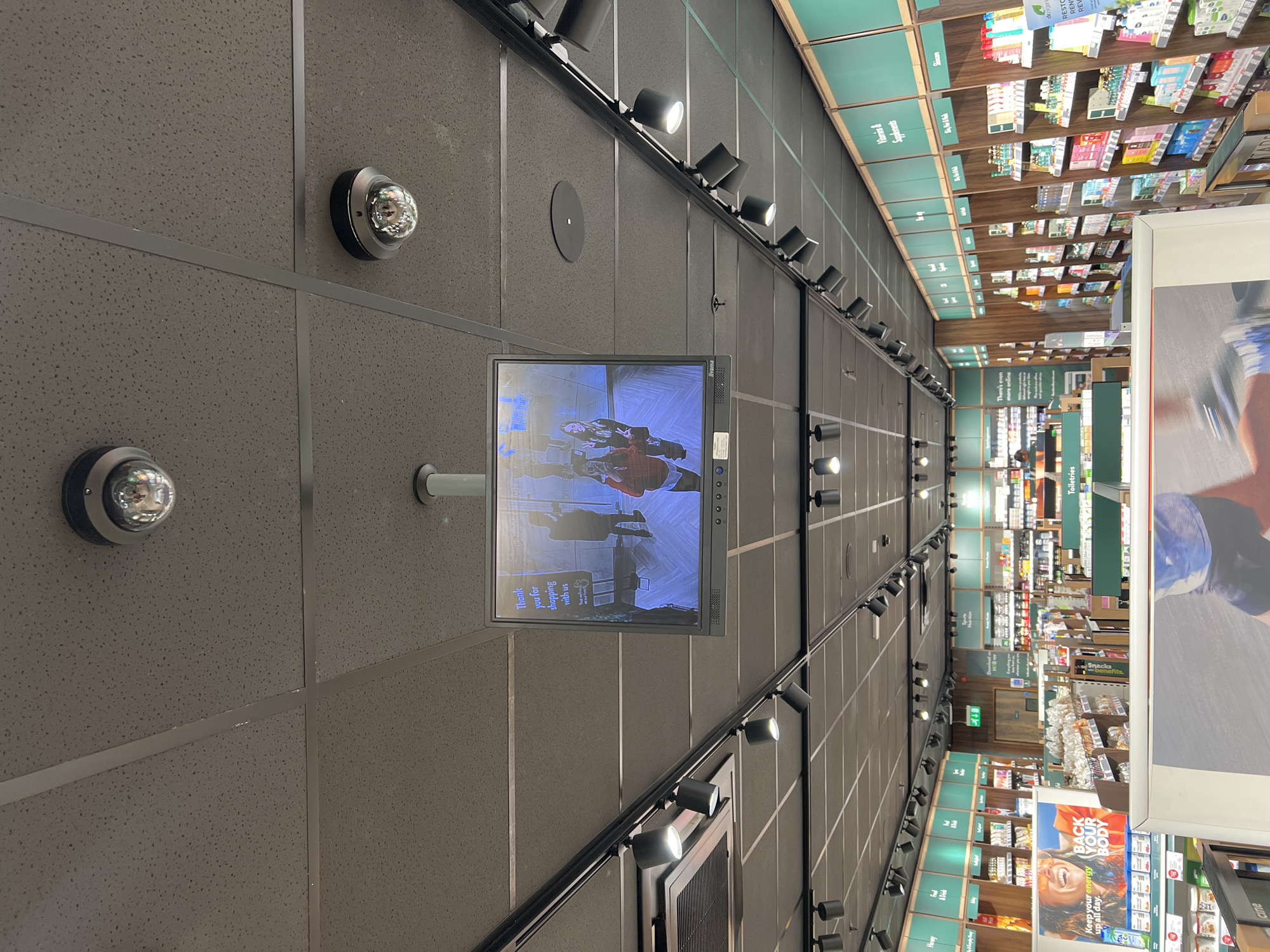 The image shows the interior of a retail store in Epsom and Ewell, United Kingdom, with a focus on the ceiling. The ceiling is gray and textured, with a grid pattern created by metal beams. Various objects are affixed to the ceiling, including dome-shaped security cameras, track lighting fixtures, and a centrally positioned television monitor displaying store security footage. The lower portion of the frame shows a section of store shelves containing products. A large advertisement is also visible, prominently featuring the phrases "BACK YOUR BODY," "Keep your energy up all day," and "cine" (possibly advertising the brand or store).