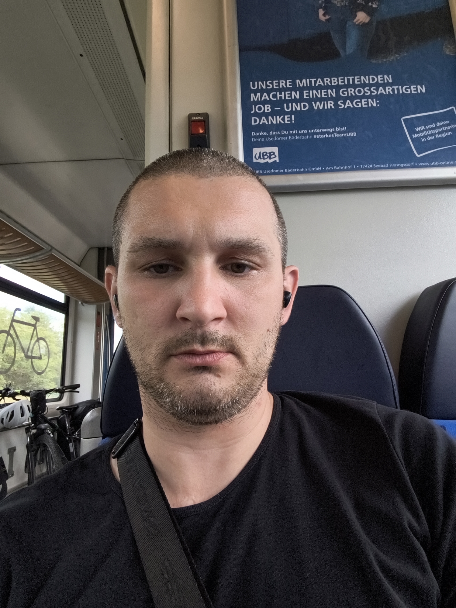 A man is taking a selfie inside a train compartment. He is wearing a black t-shirt and has short hair with a goatee. He is also wearing wireless earbuds. Through the window behind him, two bicycles can be seen. The train compartment has blue seats, and a poster is visible on the wall. The poster has text in German, which translates to "Our employees do a great job – and we say: thank you!" The train appears to be traveling on the Usedomer Bäderbahn line, serving the region of Züssow, Germany.