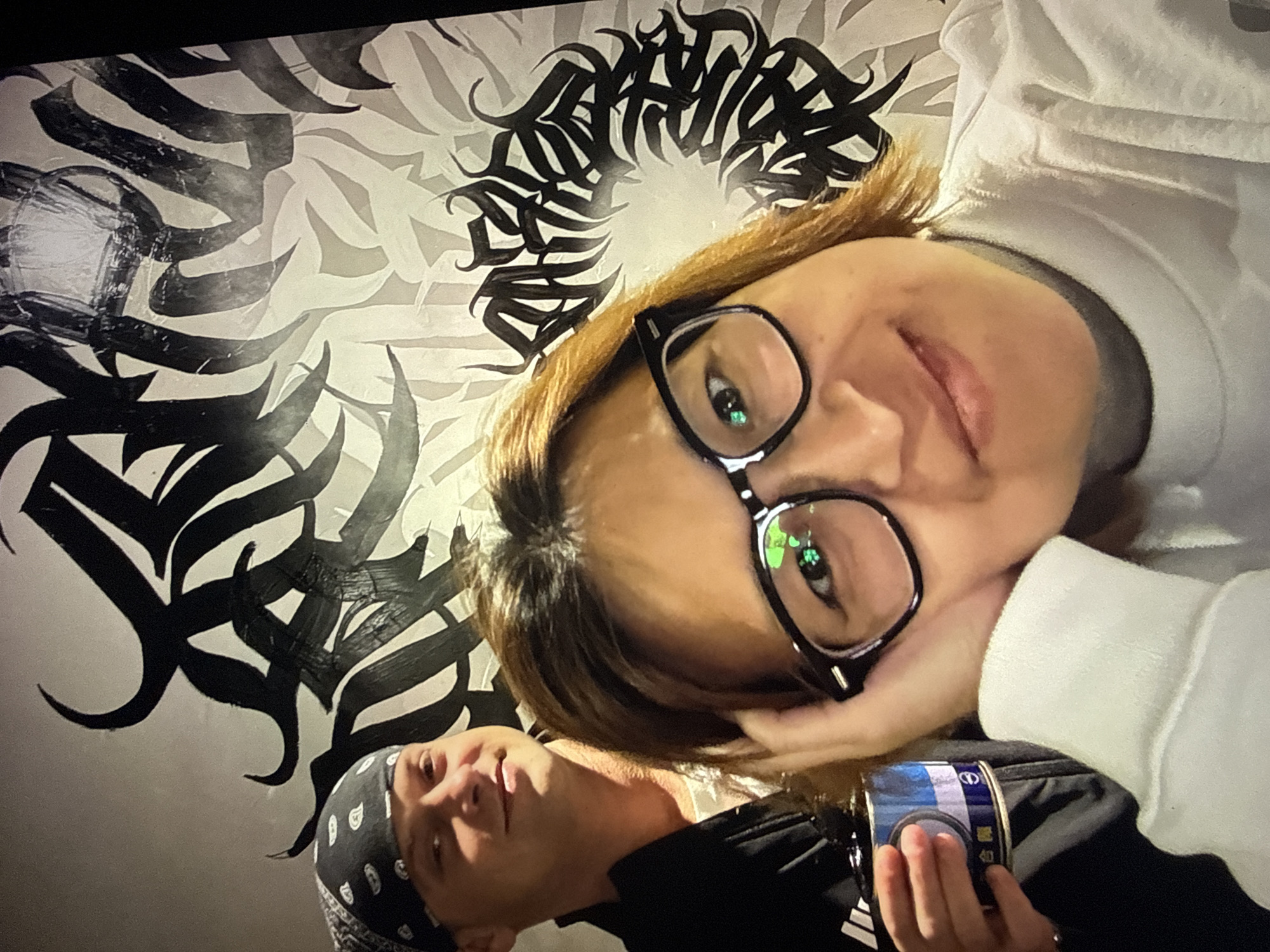 Two individuals are in the foreground, posing for a selfie in front of a wall with black and white calligraphic art. The person in the foreground on the right is a woman with shoulder-length auburn hair, wearing black-rimmed glasses and a white sweatshirt. She is resting her right hand on her head, and her left hand is partially visible. The person in the background on the left is a man with fair skin and short, light brown hair, wearing a black bandana and a black jacket over a white t-shirt. He is holding a can with blue and white labeling, which appears to be a food or beverage can, in his left hand. The wall behind them features abstract, swirling black brushstrokes on a white background, with some areas of lighter grey. The overall lighting is warm and appears to be from an indoor source. The location is not explicitly identifiable as Taipei, Taiwan.