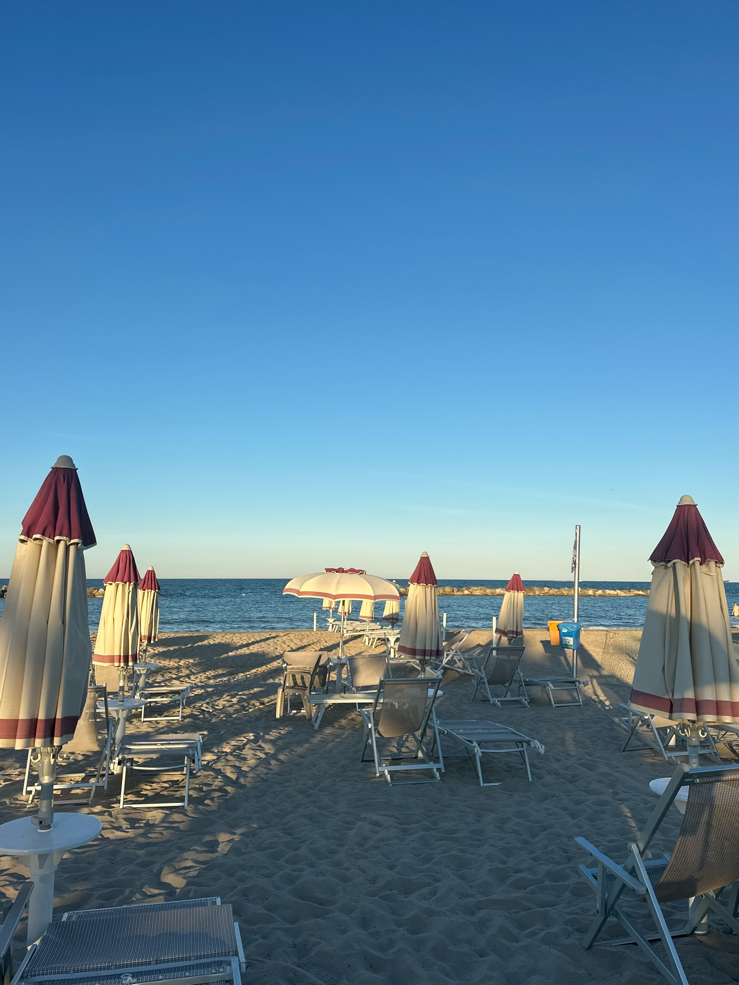The image depicts a beach scene in Civitanova Marche, Italy, with numerous closed umbrellas and lounge chairs set up on the sand. The sky is clear and blue, and the sea is calm. In the foreground, several closed beach umbrellas with burgundy and cream stripes stand tall, casting long shadows across the sand. Lounge chairs, some made of metal and fabric and others of white plastic, are arranged in rows. A small white table is visible on the left. In the mid-ground, the sea stretches to the horizon, with a rocky breakwater separating the beach from the open water. A few more umbrellas and chairs are visible further down the beach. In the background, the coastline of Civitanova Marche can be seen. The scene appears to be set in the early morning or late afternoon, given the angle of the sun and the long shadows. No people are visible in the image.
