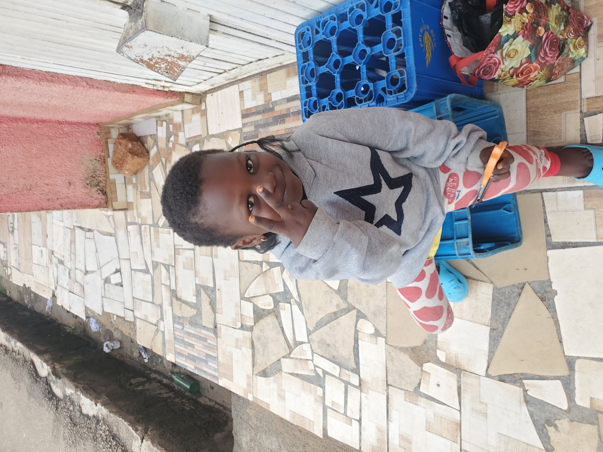 A young Black girl with braided hair is sitting on a tiled walkway in Jos, Nigeria. She is wearing a grey hooded sweatshirt with a black star design and pink patterned leggings. She is holding an orange tool and making a peace sign with her hand. In the background, there are blue plastic crates, a decorative bag with floral patterns, a pink wall, and a white door. A drainage ditch is visible to the left of the walkway, with some litter in it.