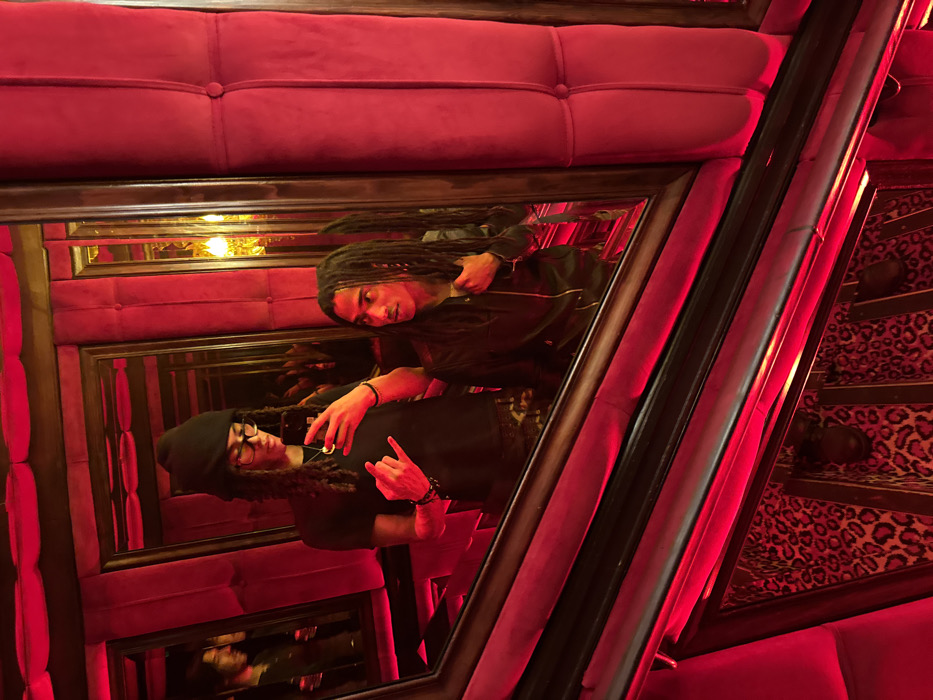 The image depicts two individuals within an interior venue located in Paris, France, characterized by an abundance of mirrors, red velvet upholstery, and pervasive red ambient lighting.
One individual, positioned on the left, is taking a selfie using a dark-colored smartphone held in their right hand. This person wears a black beanie, glasses, a black t-shirt, and has long dreadlocks or braids. Their left hand is raised, forming a "rock on" gesture. A necklace with a round pendant and bracelets are visible.
The second individual, positioned on the right, wears a dark, zippered jacket and also has long dreadlocks or braids. This person is looking towards the right, slightly upward, with their right hand near the jacket's lapel. An earring is visible in their right ear.
The environment is dominated by multiple framed mirrors with dark, possibly wooden or metallic, frames, creating a strong sense of depth and numerous reflections of the subjects and the room. The walls feature prominent red velvet paneling, some appearing tufted. The entire scene is bathed in a distinct red light, originating from unseen sources. In the deeper reflections, a warm, yellow-orange light fixture is discernable. The lower section of the image shows a dark railing, likely part of a staircase, situated above a floor or lower wall covered in a leopard print pattern. The decor suggests an entertainment venue such as a bar, club, or cabaret.