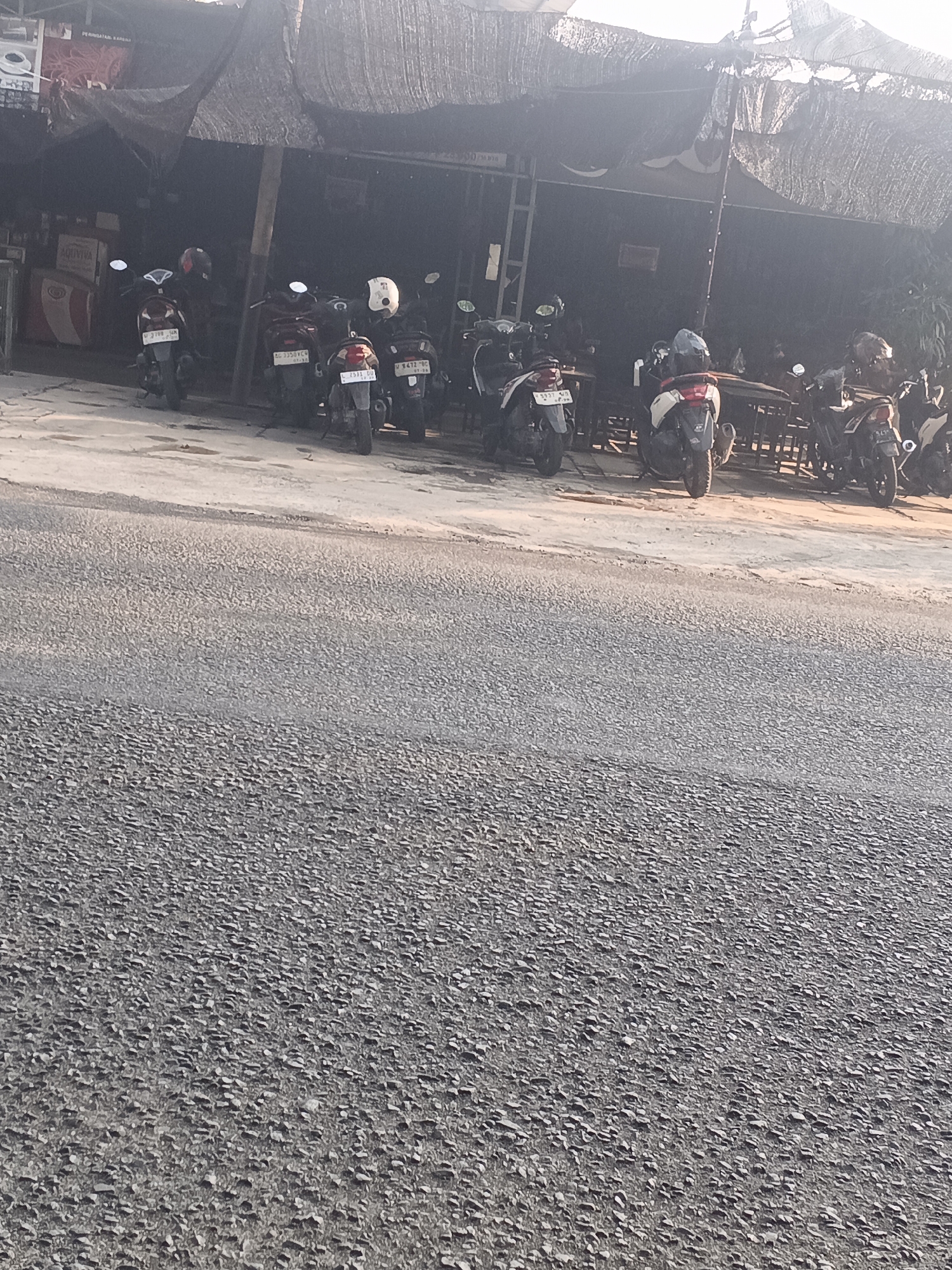 In Kemangsen, Indonesia, a row of parked motorcycles is visible in front of a low-rise building with a shaded awning. The motorcycles, mostly scooters, are positioned parallel to the building's facade. The ground in front of the building appears to be a dirt or gravel surface, transitioning into a paved road in the foreground. The building itself has an open-air structure, with some tables and chairs visible inside. The lighting suggests it is daytime, with shadows cast by the awning and the motorcycles. No people are actively engaged in any interactions within the frame.