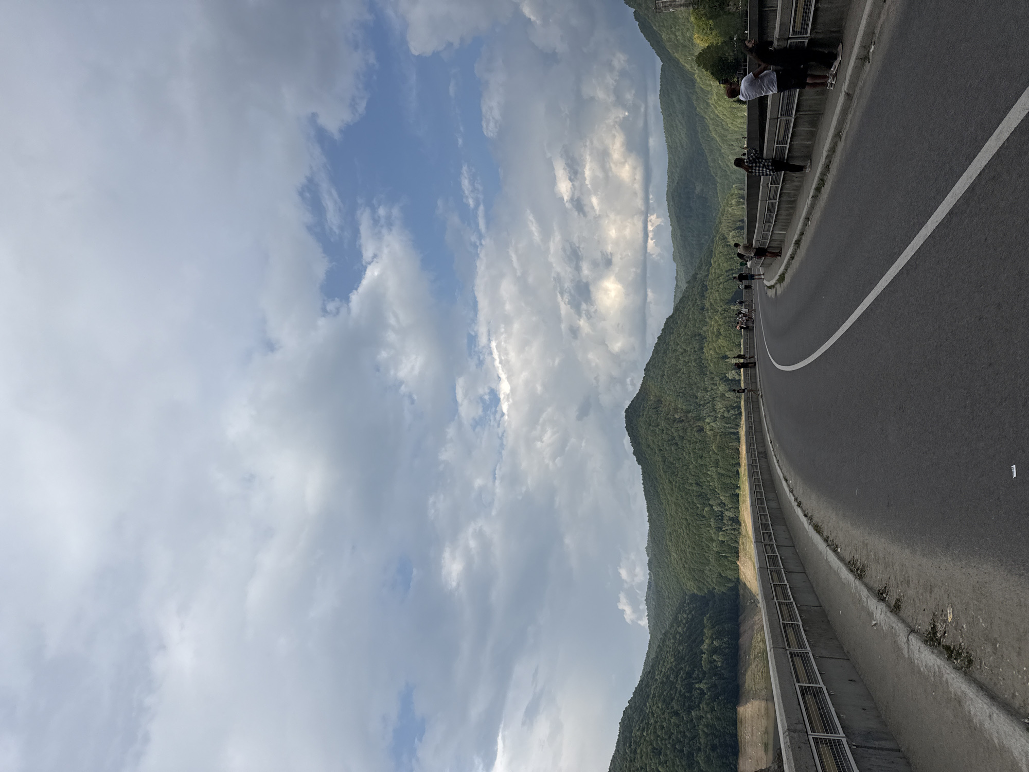 The image shows a paved road with a white line marking the center, curving to the right. The road is bordered by a concrete barrier with metal railings. On the right side of the road, several people are standing, looking out over a valley. On the left side of the road, the terrain slopes down to a body of water, with a dry, stepped bank visible. Beyond the water, lush green hills covered in trees rise up, forming a valley. The sky above is filled with dramatic, cloudy formations, with patches of blue sky visible. The scene is set in a mountainous, natural environment. The context provided indicates this location is in Arefu, Romania, likely a dam or a viewpoint overlooking a reservoir and surrounding landscape.