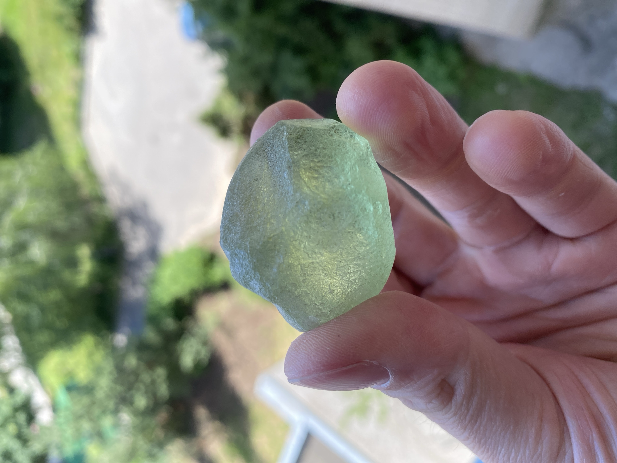 In this close-up shot, a person's hand holds a translucent, pale green, irregularly shaped object, possibly a piece of sea glass or a rough gem. The fingers, with visible lines and texture, gently grip the object, showcasing its frosted surface and slightly angular facets. The background is softly blurred, revealing hints of a natural, outdoor setting. Lush green foliage and the washed-out tones of what appears to be a pathway or ground suggest a daytime scene, likely under natural light. The overall impression is one of casual observation, perhaps of a found natural item. The setting hints at a possibly residential or park-like area, given the greenery and hints of structures. There is no visible text in the image.