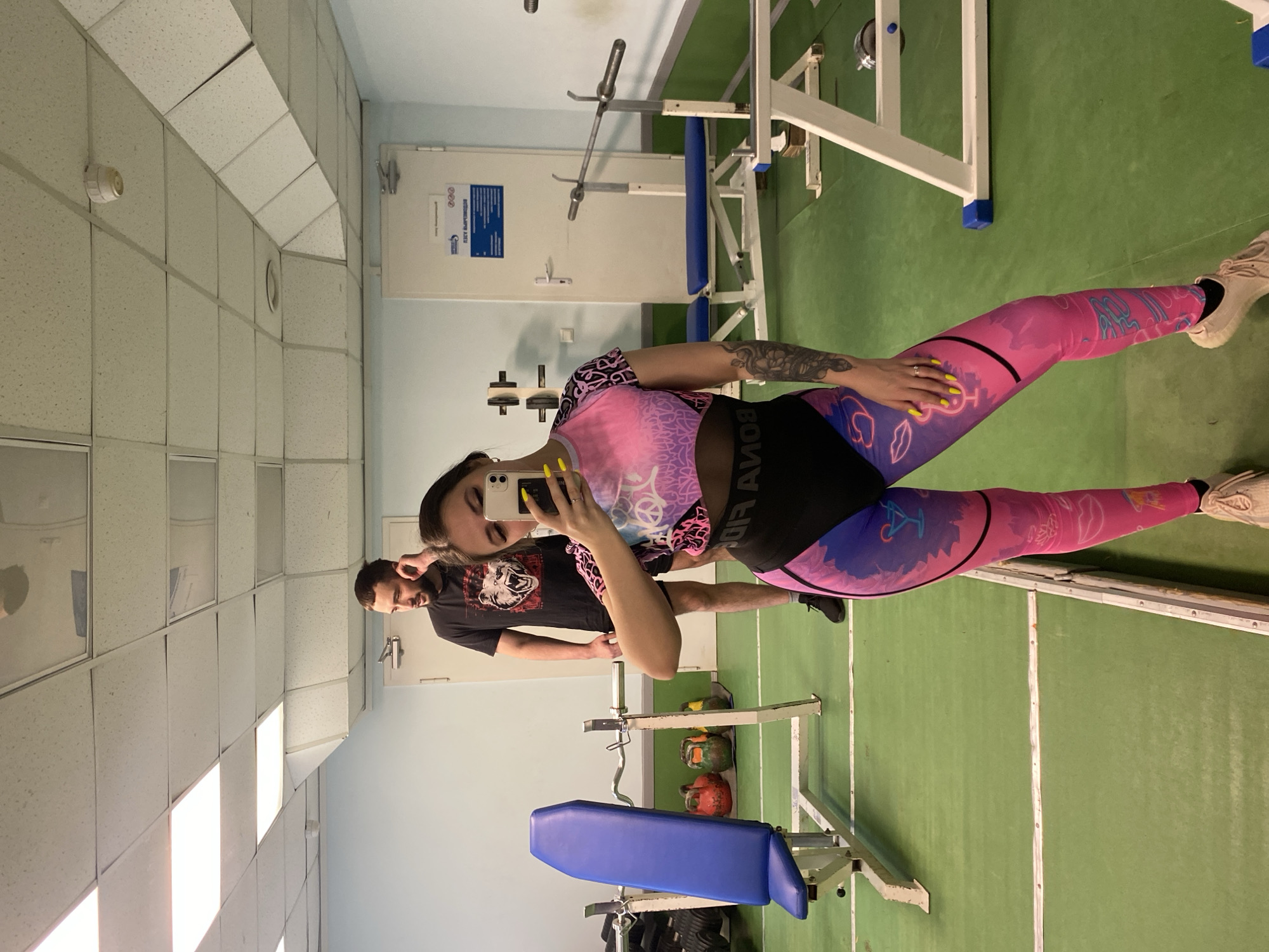 The image depicts a woman taking a selfie in what appears to be a gym. She is wearing a pink and purple patterned athletic outfit and has a visible tattoo on her arm. Behind her stands a man wearing a black t-shirt. The gym contains standard weightlifting equipment, including a weight bench and kettlebells. The room has a green floor, white walls, and fluorescent lighting. The location is potentially in Chaikovsky, Russia, though this is unconfirmed based on visual information alone.