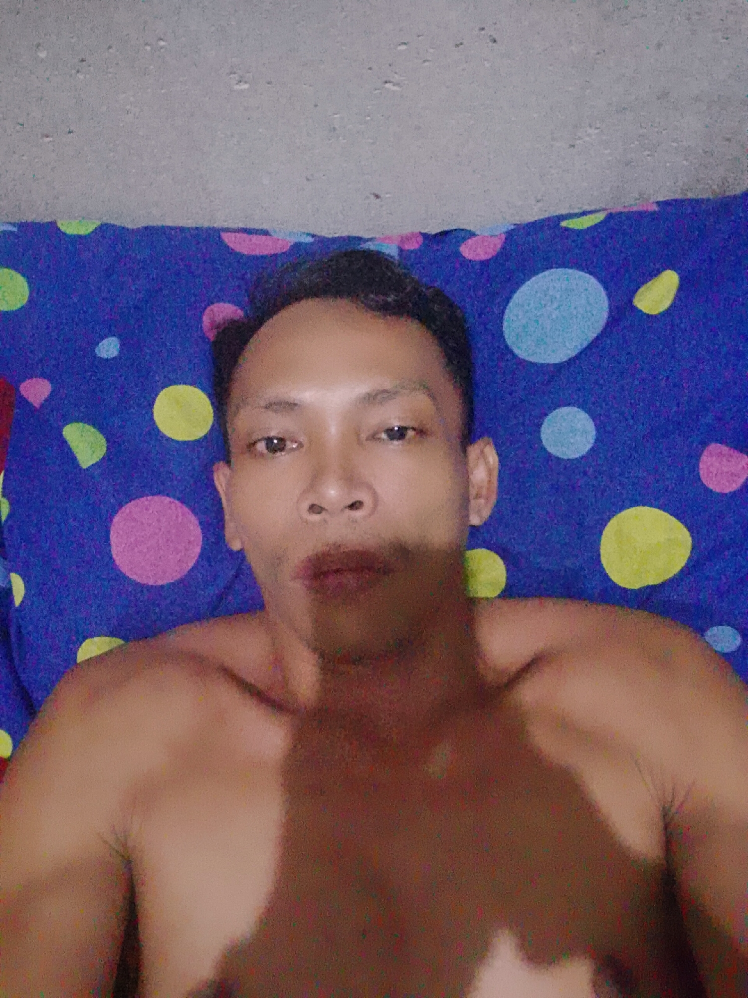 A shirtless person lies on a blue pillow with multicolored polka dots. The person is facing the camera. A gray concrete ceiling is visible in the background. The location is Bukit Ketok, Indonesia.