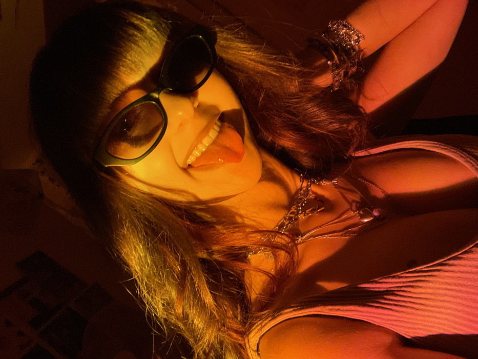 A woman with long brown hair and sunglasses is sticking out her tongue. She is wearing a pink ribbed top and several silver necklaces. Multiple silver bracelets adorn her wrist. The image is bathed in warm, orange lighting, creating a vibrant, almost surreal effect.