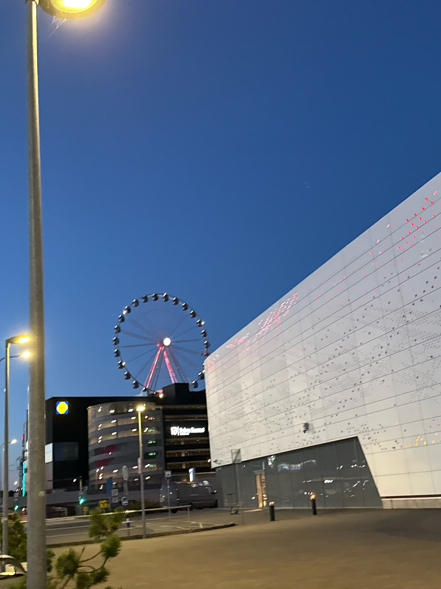 In this image, a wide-angle shot captures a twilight scene in Tallinn, Estonia. The sky is a deep, clear blue, indicating it is likely evening or early morning. Dominating the right side of the frame is a large, modern white building with a distinctive facade. This building is adorned with a pattern of small, irregularly spaced red lights, suggesting it might be an entertainment venue or a modern architectural structure.
To the left of the white building, a Ferris wheel stands prominently against the sky, its circular structure outlined by numerous lights, with a bright pink glow emanating from its center and spokes. The Ferris wheel is situated behind a cluster of buildings, including a dark rectangular building with a bright yellow circular logo, identified as Lidl. Adjacent to the Lidl building is a rounded, multi-story structure with illuminated windows.
In the foreground, a tall, silver lamppost with a bright yellow light fixture is visible on the left. Another lamppost with a similar yellow light is positioned further back. The ground in the foreground appears to be a paved area, possibly a street or a pedestrian walkway. There are also hints of greenery, with small pine trees visible in the lower left corner. The overall impression is a blend of urban infrastructure, modern architecture, and recreational elements within the city.