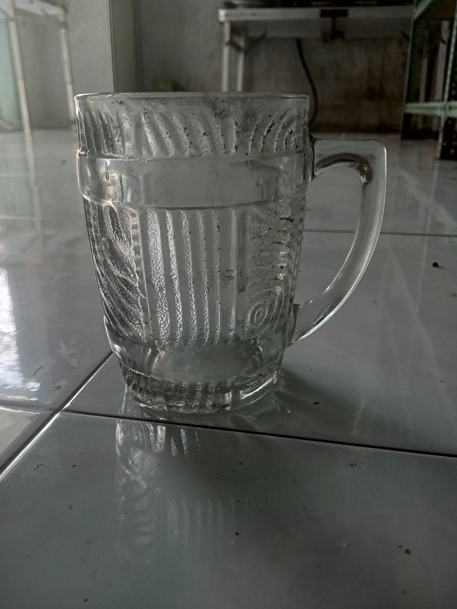A clear glass mug with a textured pattern of vertical lines and curved leaf-like designs sits on a tiled floor. The mug is empty and reflects the surrounding environment. The floor tiles are light gray and have a slight sheen. In the background, a blurred view shows a wall, a table or counter, and some shelving. The overall scene appears to be indoors, likely in a domestic or commercial setting. The image does not contain any people or specific indicators of the city of Badas, Indonesia, beyond the initial prompt.