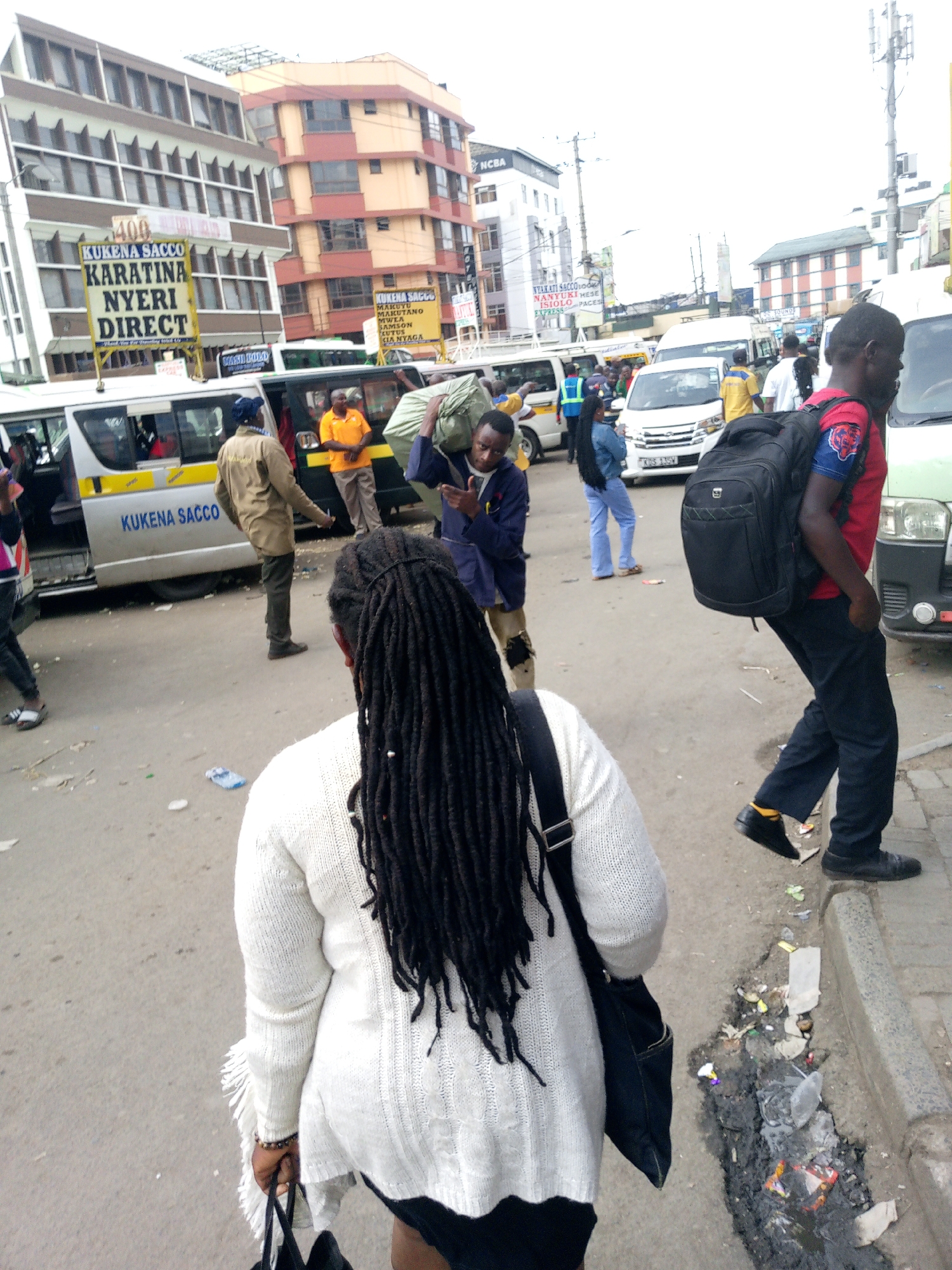 The image depicts a bustling street scene in Nairobi, Kenya. A woman with long dreadlocks and wearing a white sweater and black skirt is walking away from the camera. Several people are around buses, which are identified as "Kukena Sacco" buses with destinations listed as "Karatina Nyeri Direct." Other destinations are listed on nearby signage. A man in a red shirt and black pants is walking along the curb, carrying a black backpack. Litter and debris are visible in the street and along the curb. Buildings with signs such as "NCBA" and "Nyakati Sacco" are visible in the background. People are either boarding or disembarking from the buses.