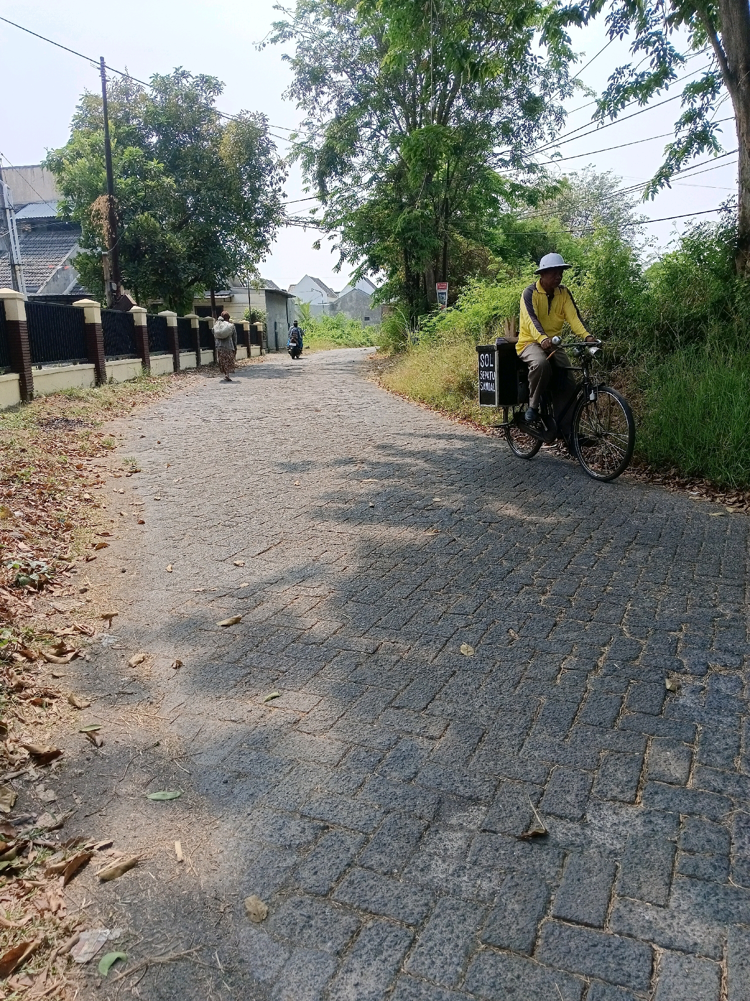 A man wearing a yellow shirt and a light-colored hat rides a bicycle on a paved road in Tawangsari, Indonesia. The bicycle has a black box attached to the back, with white lettering that reads "SOL SEPATU SANDAL." The road is constructed of dark gray interlocking pavers and is lined with dry leaves and grass on the left side. On the right side of the road, there is dense green vegetation. In the background, there are houses with tiled roofs and fences, as well as trees and power lines. A person wearing a patterned skirt and carrying a bundle on their shoulder walks away from the camera in the distance, and another person rides a scooter. The sky is bright and overcast.