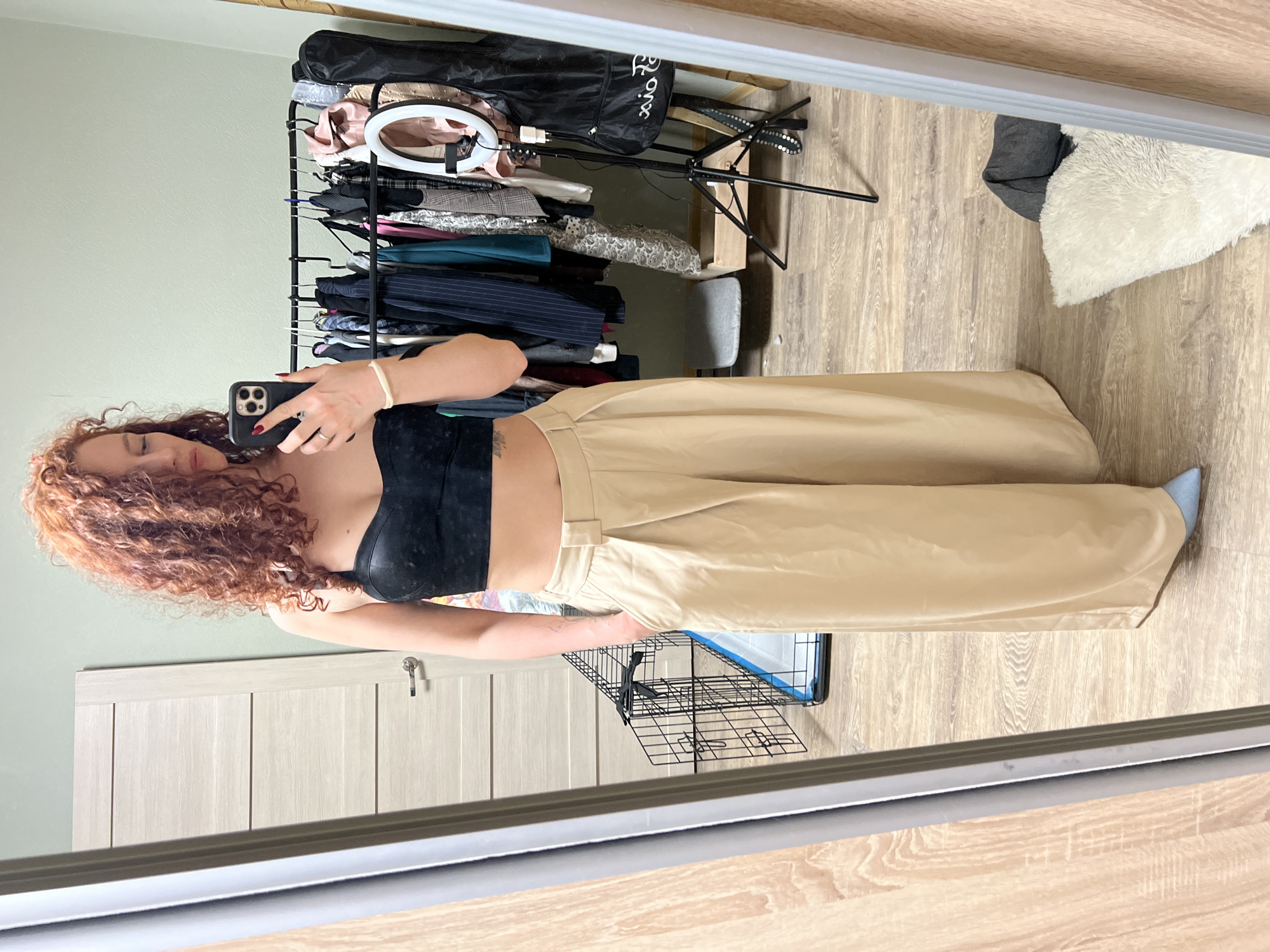 The image displays a mirror selfie of a woman with curly red hair, taken in an indoor setting in Saint Petersburg, Russia. The woman is wearing a black, strapless crop top and loose-fitting, light beige wide-leg pants. She holds a dark-colored smartphone with three camera lenses up to take the photo, with her eyes closed or looking down. Her right hand has dark nail polish and a ring on her ring finger, while her left wrist sports a light-colored bracelet and her forearm shows a visible tattoo. A light blue shoe or slipper peeks out from under her right pant leg.
The room, as reflected in the mirror, features light wood-laminate flooring and walls painted a light green or grey hue. To the left of the woman is a light-colored wooden door with vertical paneling. To her right, a black metal clothing rack is laden with various garments, including jackets, blouses, and pants in shades of black, grey, blue, and patterned fabrics. A white ring light with a stand is positioned near the top of the clothing rack, and a black bag with the text "xo Pa" is visible resting on the rack. Below the clothing rack, a small grey stool or pouf is visible, along with a black wire basket. Further to the right, a white fluffy cushion or rug lies on the floor. The image appears to be taken during the daytime.