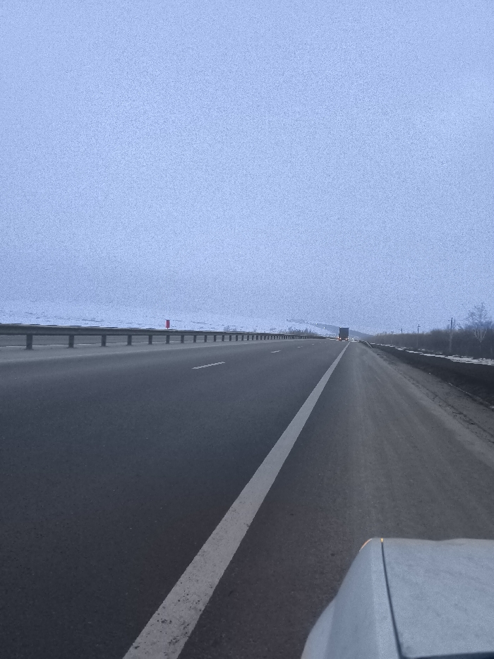 The image depicts a multi-lane asphalt highway scene, viewed from the perspective of a vehicle's dashboard or hood, in what is identified as the vicinity of Pobeda, Russia. The road surface is dark grey, with a prominent solid white line delineating the right edge of the immediate lane, extending into the background. To the left of the road, a metal guardrail runs parallel, separating the highway from snow-covered, undulating terrain that extends to the horizon. A single red, vertical marker is visible near the guardrail in the mid-distance on the left. To the right of the highway, the shoulder appears darker and less snow-covered, transitioning into sparse, bare trees and brush partially visible along the roadside. In the far distance, a semi-truck is visible, traveling in the same direction, with its red taillights illuminated. The sky occupies the upper half of the frame, presenting a uniform, textured light blue-grey, indicating heavy overcast conditions. No people are visible. The overall environment is consistent with a cold, winter day.