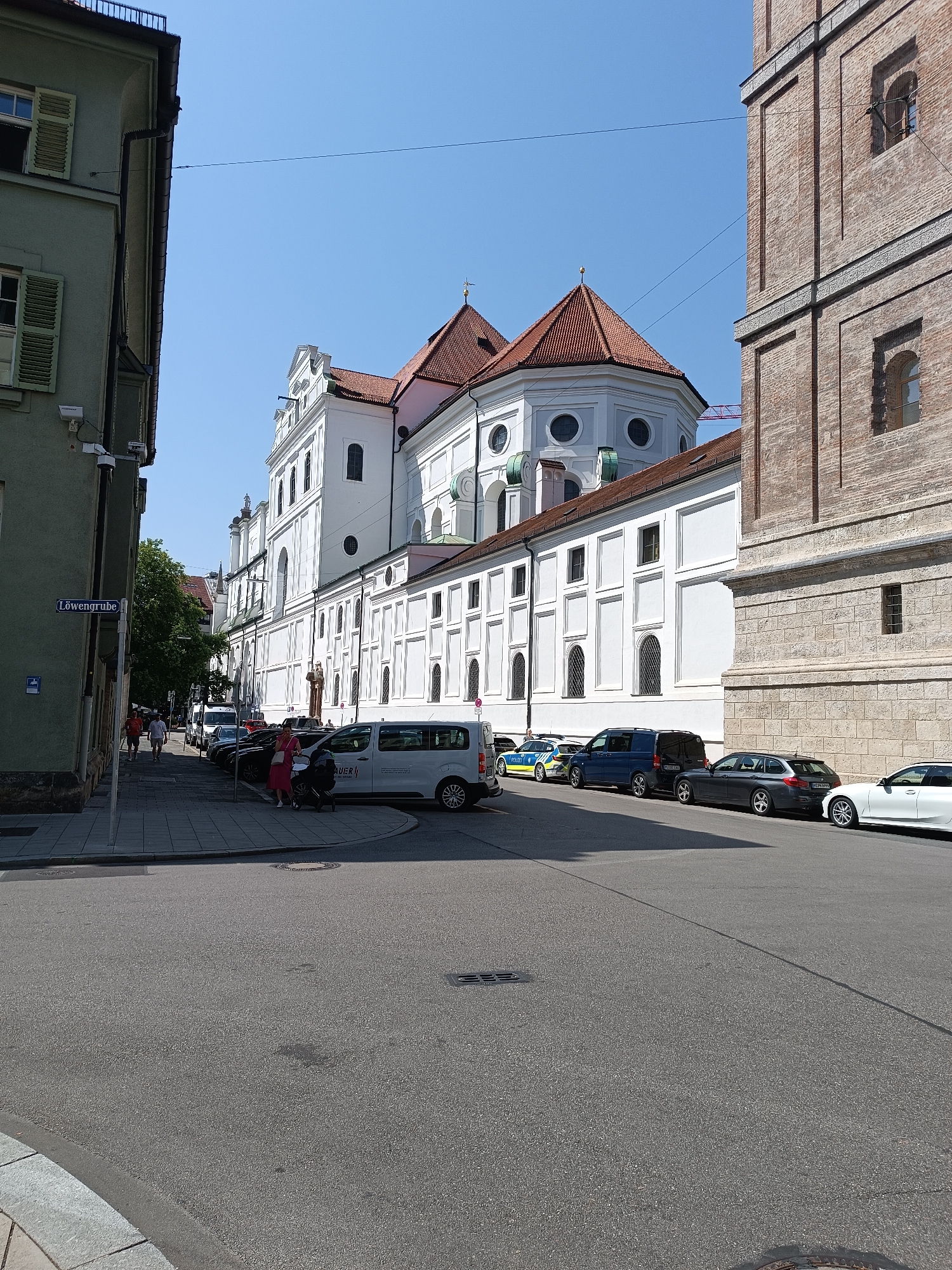 A sunny day scene depicts a street in Munich, Germany, with a large white building featuring a tiled red roof in the background. The building has multiple windows and decorative elements. To the right, a brick building with arched windows stands tall. The street is paved and lined with parked cars, including a white van with "AUER" written on it, a police car, a dark blue van, a dark gray station wagon, and a white sedan. People are seen walking on the sidewalk. A street sign labeled "Löwengrube" is visible on the left side of the image. The sky is clear and blue.