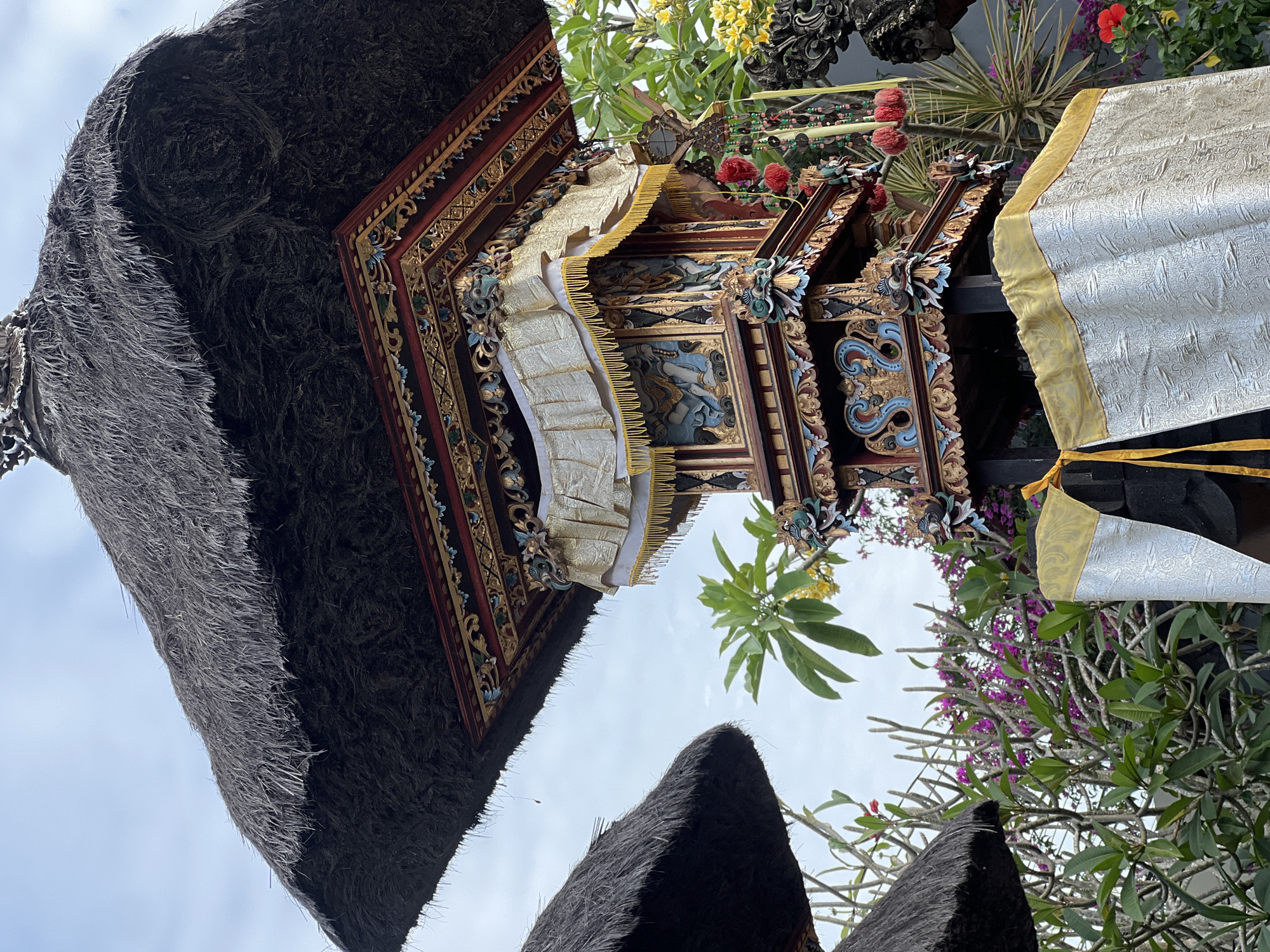 In the city of North Kuta, Indonesia, an ornate Balinese shrine stands amidst lush greenery and a cloudy sky. The shrine is multi-tiered, with a thatched roof and intricate carvings, adorned with gold fabric and decorative elements. The shrine's detailed decorations include figures within panels, decorative trim, and hanging ornaments. Surrounding the shrine are various plants, including flowering bushes with vibrant purple and red blooms, and broad-leafed green plants. The scene suggests a cultural or religious setting, possibly a temple or a ceremonial space. There are no people visible in the image.