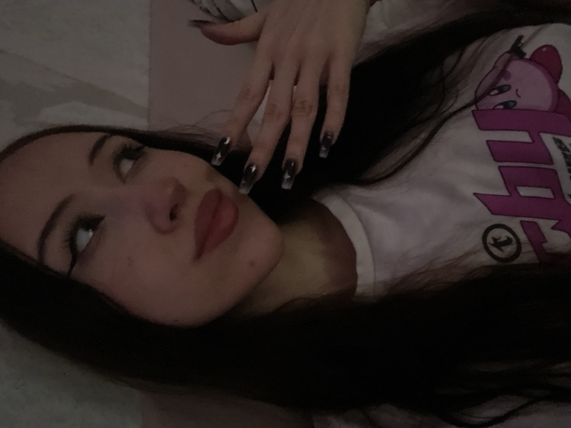 The image shows a close-up of a young woman with dark hair, looking to her right. She is wearing a white t-shirt with a pink graphic featuring a character that resembles Kirby. Her hand is raised, showing long, dark-colored fingernails. The lighting is dim, suggesting an indoor setting. The background is blurred and appears to be a wall with some textured pattern. There is no indication of Votkinsk, Russia, or any specific city in the image.
