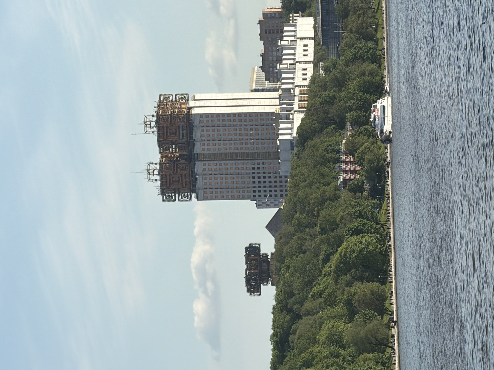 The image shows a view of the Russian Academy of Sciences building in Moscow, Russia, along the Moskva River. The building, a high-rise structure with a distinctive, elaborate superstructure, stands prominently against a blue sky with scattered clouds. To the right of the building, a dense green expanse of trees lines the riverbank. Along the water's edge, a pathway is visible with people walking. Several boats are docked along the river. The river itself is a dark, rippling body of water, filling a significant portion of the right side of the image.