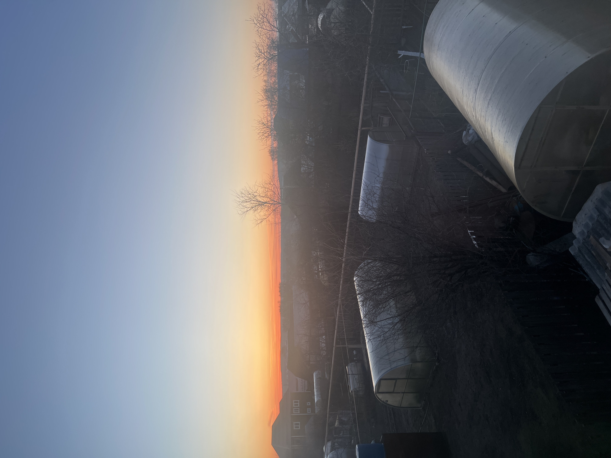 The image shows a view of Votkinsk, Russia at dawn or dusk. The sky transitions from a clear blue to orange and red near the horizon, indicating the sun's position. In the foreground, there are two large, cylindrical structures, likely tanks or silos, which are casting shadows. There are also what appear to be two greenhouses. Leafless trees dot the scene, suggesting it's late fall or winter. In the background, there are residential buildings, some with visible windows. There appears to be some fog or haze present, obscuring the distant details of the city. There are also some metal beams stacked at the bottom right corner.