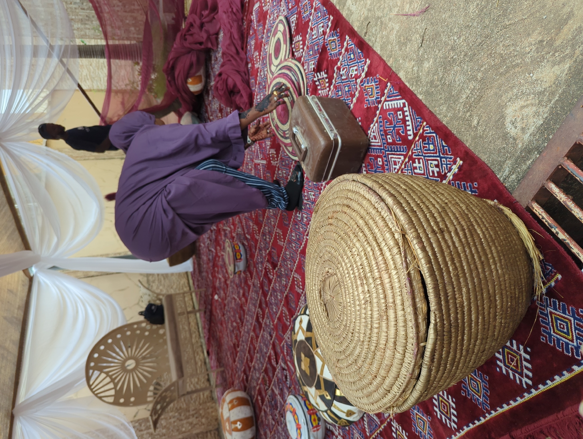 The image captures a vibrant cultural setting, likely an outdoor or semi-outdoor event in Unguwar Rimi, Nigeria. The scene is dominated by a rich, red patterned rug covering much of the ground.
In the foreground, a person dressed in a long, flowing purple garment with striped trousers is seated on the rug, facing away from the camera. Their left hand, adorned with intricate henna designs and rings, is visible near some smaller, round, woven decorative mats placed on the rug. Next to them is a vintage-style brown hard-shell suitcase. To the right, a large, round, lidded basket made of woven straw occupies the immediate foreground.
The surrounding environment features decorative elements such as white and maroon/red sheer fabrics draped, suggesting a temporary canopy or tent setup. In the background, another person wearing a black t-shirt can be seen standing. A small, ornate metal table with a sunburst pattern is also visible. The area beyond the rug appears to be a concrete or dirt ground, with a metal grate visible on the far right edge.
The presence of henna, traditional woven items, and the patterned rug strongly indicates a cultural celebration or ceremony, possibly a wedding or engagement, which is common in Nigerian traditions. The image appears to be taken during daylight hours. No discernible text is visible.