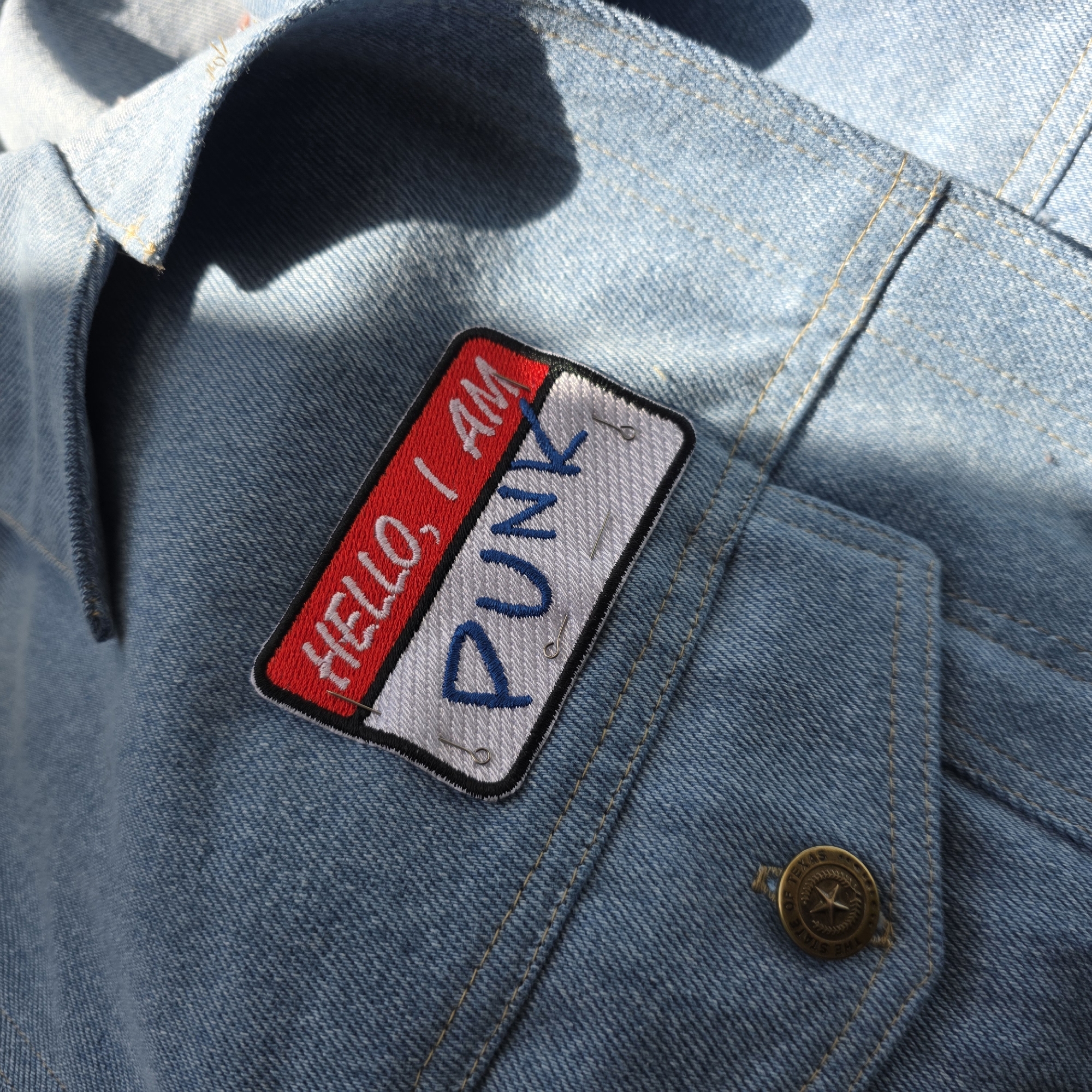 A close-up shot captures a light blue denim jacket, bathed in bright sunlight that casts sharp shadows across its textured surface. The jacket features prominent light-colored stitching and a brass-colored button visible in the lower right, embossed with "THE STATE OF TEXAS" encircling a star design.
The focal point is an embroidered rectangular patch, pinned onto the jacket with several small safety pins. The patch is divided into two sections. The left half is red with white embroidered text that reads "HELLO, I AM". The right half is white with blue embroidered text that boldly states "PUNK". The combination of the patch and the jacket suggests a personal statement or stylistic choice.
The strong contrast between light and shadow indicates a sunny daytime setting, possibly outdoors or near a window. The jacket appears to be draped or lying down. The overall scene provides a detailed view of the fabric, stitching, and decorative elements, set in Saint Petersburg, Russia.
