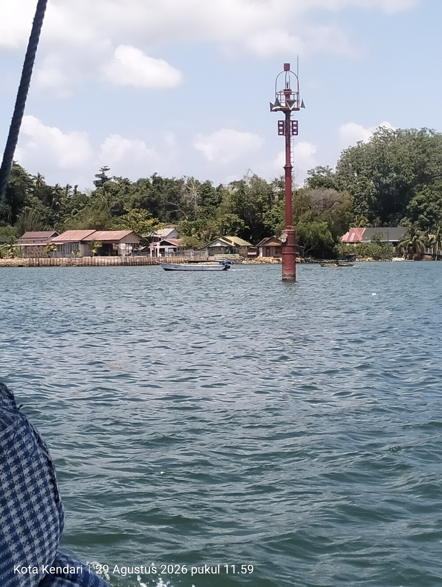 A medium-shot, eye-level, landscape photograph captures a serene daytime scene of the coastal waters in Kendari, Indonesia. The image is dominated by the calm, rippling surface of the sea in the foreground, reflecting the light blue sky dotted with white clouds. On the left side of the frame, a dark rope, possibly part of a vessel, enters from the top edge, angling downwards.
In the middle ground, a red, narrow tower stands tall in the water, topped with a cage-like structure housing what appears to be signaling equipment. This tower is positioned near a shoreline lined with a row of modest buildings, each with reddish-brown roofs. Lush green trees and vegetation densely cover the land behind the buildings, forming a backdrop of a tropical landscape.
Several small boats are visible on the water: one white boat is positioned horizontally near the shore, and a smaller, darker boat is further out in the water. The water itself is a deep blue-green, with light reflecting off the gentle waves.
The lower left corner of the image features a timestamp and location marker, indicating "Kota Kendari | 29 Agustus 2026 pukul 11.59". The overall impression is one of tranquility and a glimpse into everyday life in a coastal Indonesian city.
