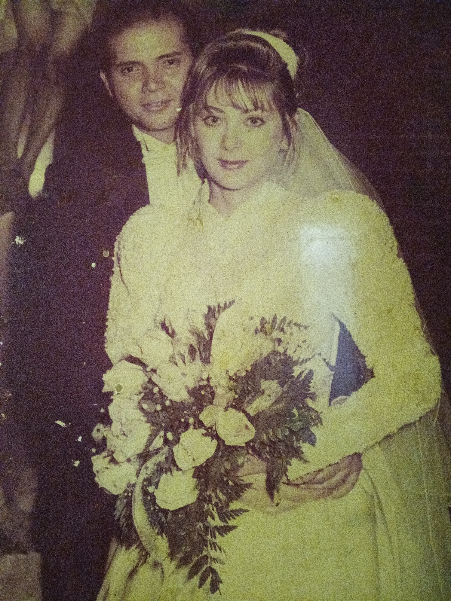 A vintage photograph shows a man and woman dressed for a wedding. The woman wears a long-sleeved white gown, a veil, and holds a bouquet of white roses. The man is behind her, wearing a dark suit with a white shirt. The location and city in Caracas, Venezuela is unable to be determined from the image.