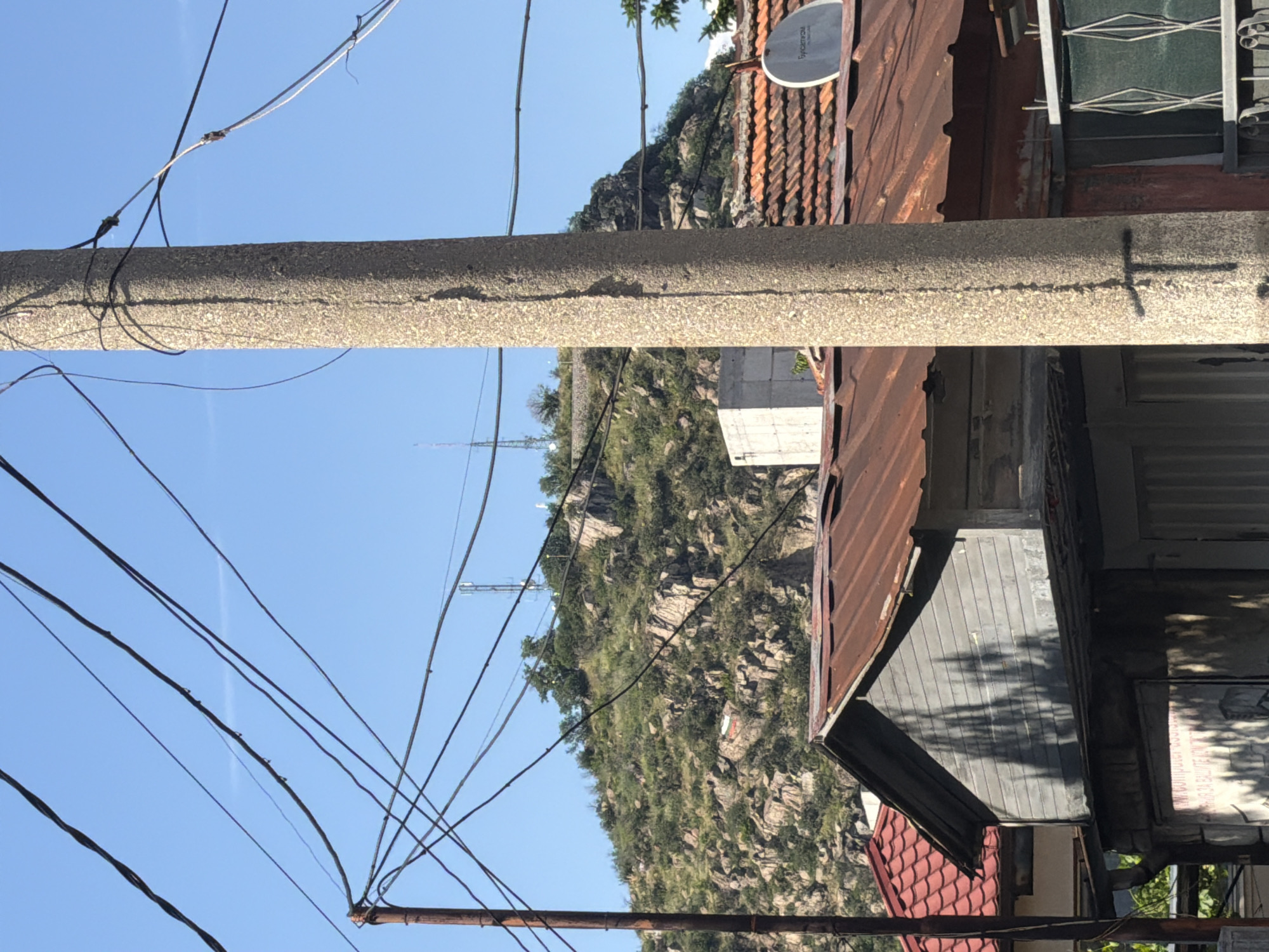 The image presents a scene in Plovdiv, Bulgaria, characterized by a juxtaposition of natural and urban elements. In the background, a rocky, verdant mountain rises, topped with what appear to be communication towers. Contrasting with this natural backdrop, the foreground is dominated by a concrete utility pole, crisscrossed by a dense network of power lines that stretch across the blue sky. To the right, a collection of weathered buildings is visible, featuring rusty corrugated metal roofs, a satellite dish, and various architectural details reflecting a modest urban environment. The scene suggests a residential area in Plovdiv situated at the intersection of urban development and natural landscapes. The objects present include a mountain, concrete pole, electrical cables, buildings and their roof.
