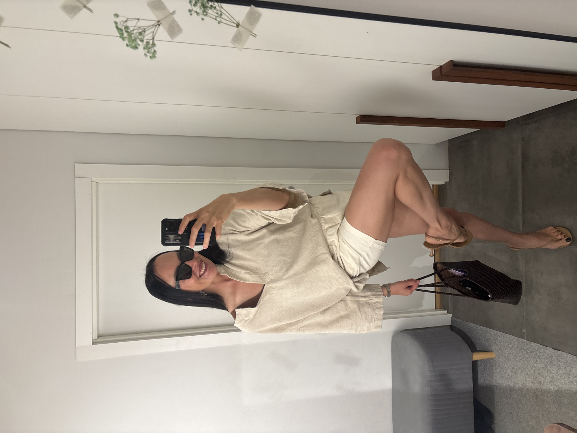 A woman is taking a selfie in a mirror. She is wearing a beige linen shirt and white shorts. She is also wearing sunglasses and sandals. She is holding a brown purse. The background is a white wall and a gray floor. There is a gray stool in the corner of the room. The location is unknown, but it is not in the city of Chaikovsky, Russia.