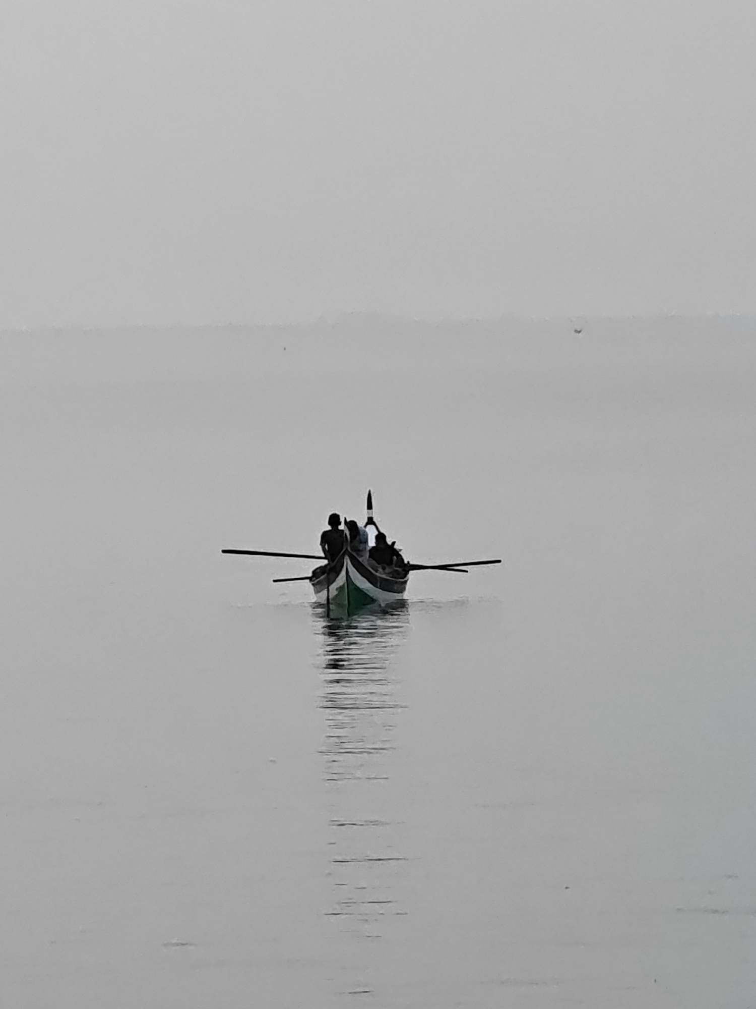 A serene and minimalist image depicts a small, traditional wooden boat carrying several individuals across calm, misty waters in Lhokseumawe, Indonesia. The boat, positioned centrally, features a distinct green stripe along its hull, contrasting with darker tones. At least three people are visible within the boat as dark silhouettes: one at the bow, one in the middle, and another towards the stern. Oars or paddles extend from both sides of the vessel, indicating active rowing or paddling.
The entire scene is enveloped in a soft, diffused gray light, strongly suggesting a misty, foggy, or heavily overcast day, possibly during early morning or late afternoon. This atmospheric condition blurs the horizon line, creating a seamless blend between the tranquil water and the sky. The water's surface is remarkably still, producing a clear yet subtly rippled reflection of the boat and its occupants. No other boats, land features, or structures are visible in the expansive frame, enhancing the sense of isolation and quietude. The overall impression is one of peaceful solitude and a harmonious interaction with nature. There is no visible text in the media file.
