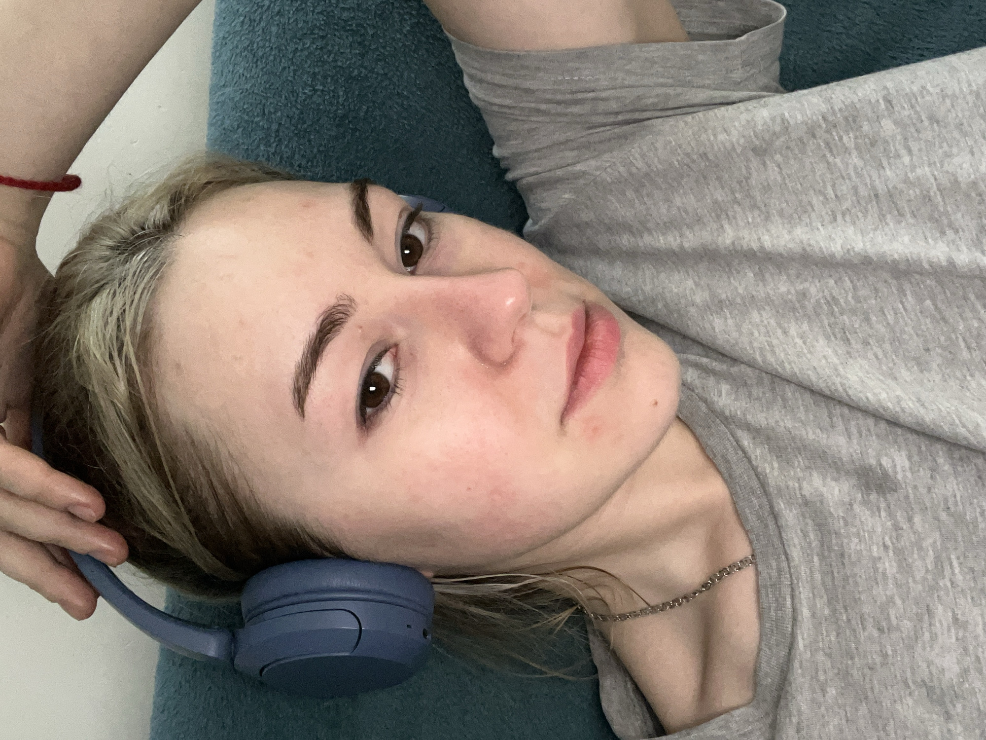 A young woman is lying down, possibly on a bed or similar surface. She has blonde hair with brown eyebrows and eyes. She is wearing a gray shirt and a silver necklace. She is holding blue headphones against her left ear. Her left arm is visible above her head and a red bracelet is around her wrist. The background is a teal or blue textured surface. There is no evidence to suggest the location is Boreti, Montenegro.