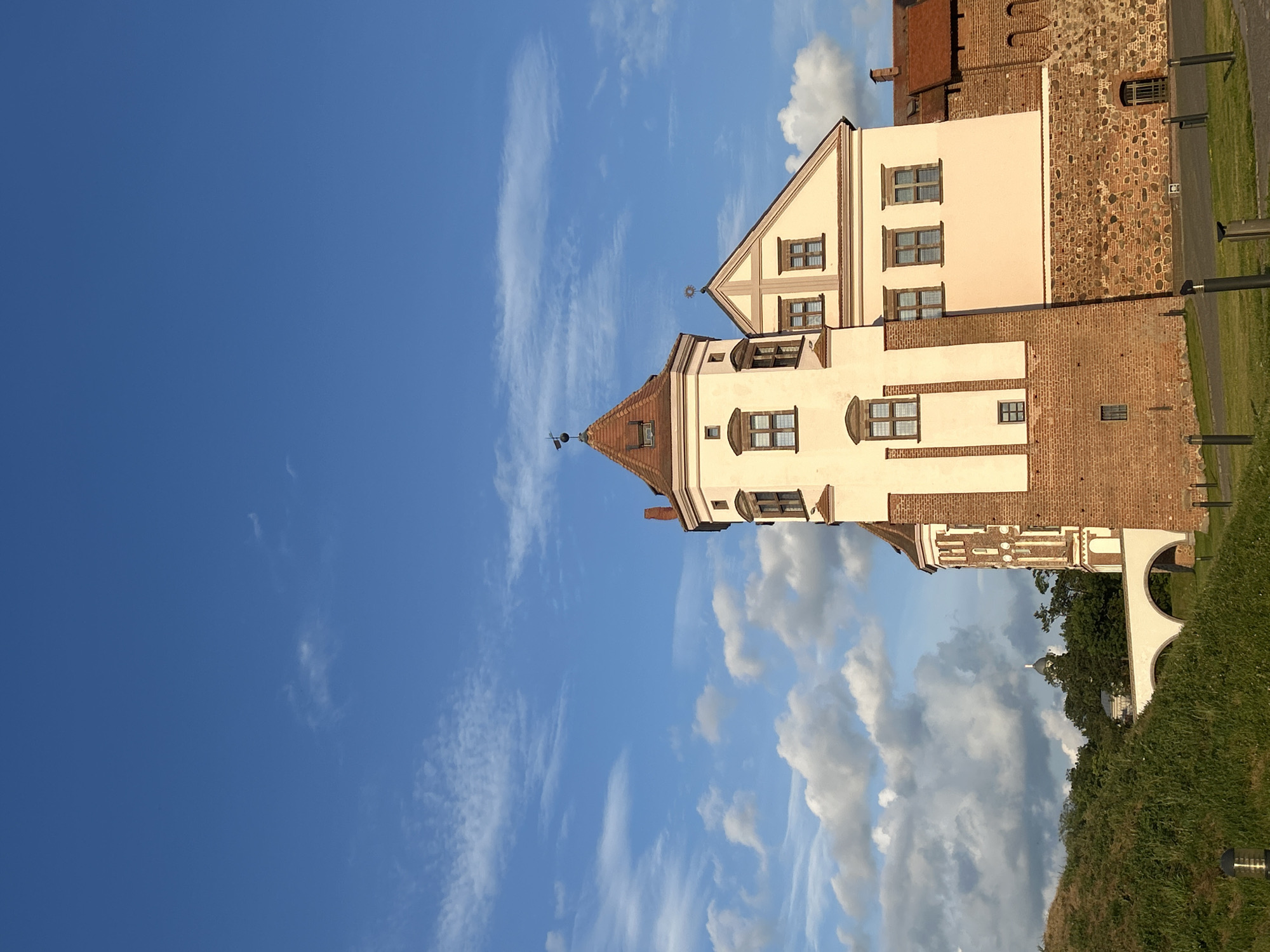 This is a vertical, eye-level shot of a tower on a castle with a bright blue sky with scattered white clouds overhead. The tower is made of red brick with tan, stucco-like walls. It has several windows, some of which are arched, and a red-tiled roof topped by a weather vane. To the right of the tower is a smaller, peaked roof section with a cross and a sunburst emblem. To the left of the tower, a grassy mound slopes down towards a white arched bridge. In the distance, behind trees, is a building with a dome. The ground in front of the castle is paved with stones and grass, with several black bollard-like lights. The scene appears to be set in the village of Mir, located in the Zhukavichy Selsoviet of Belarus.