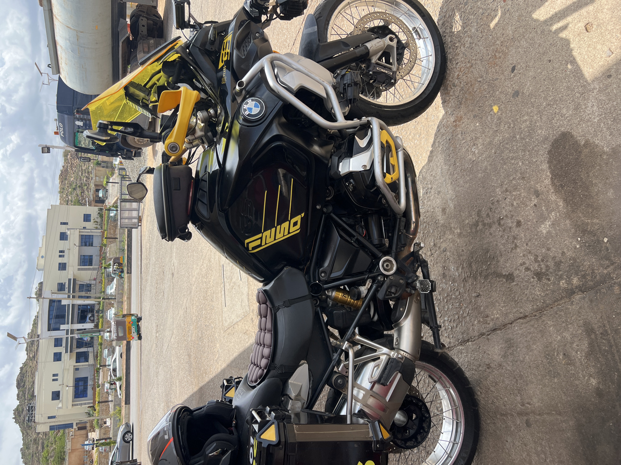 The image shows a black BMW motorcycle parked on a concrete surface, likely a parking lot or street. The motorcycle has yellow accents and features a crash bar. In the background, there are buildings, trees, and a tricycle on the road, suggesting an urban setting. There is also a large tanker truck behind the motorcycle. The location is Gura Top, Nigeria.